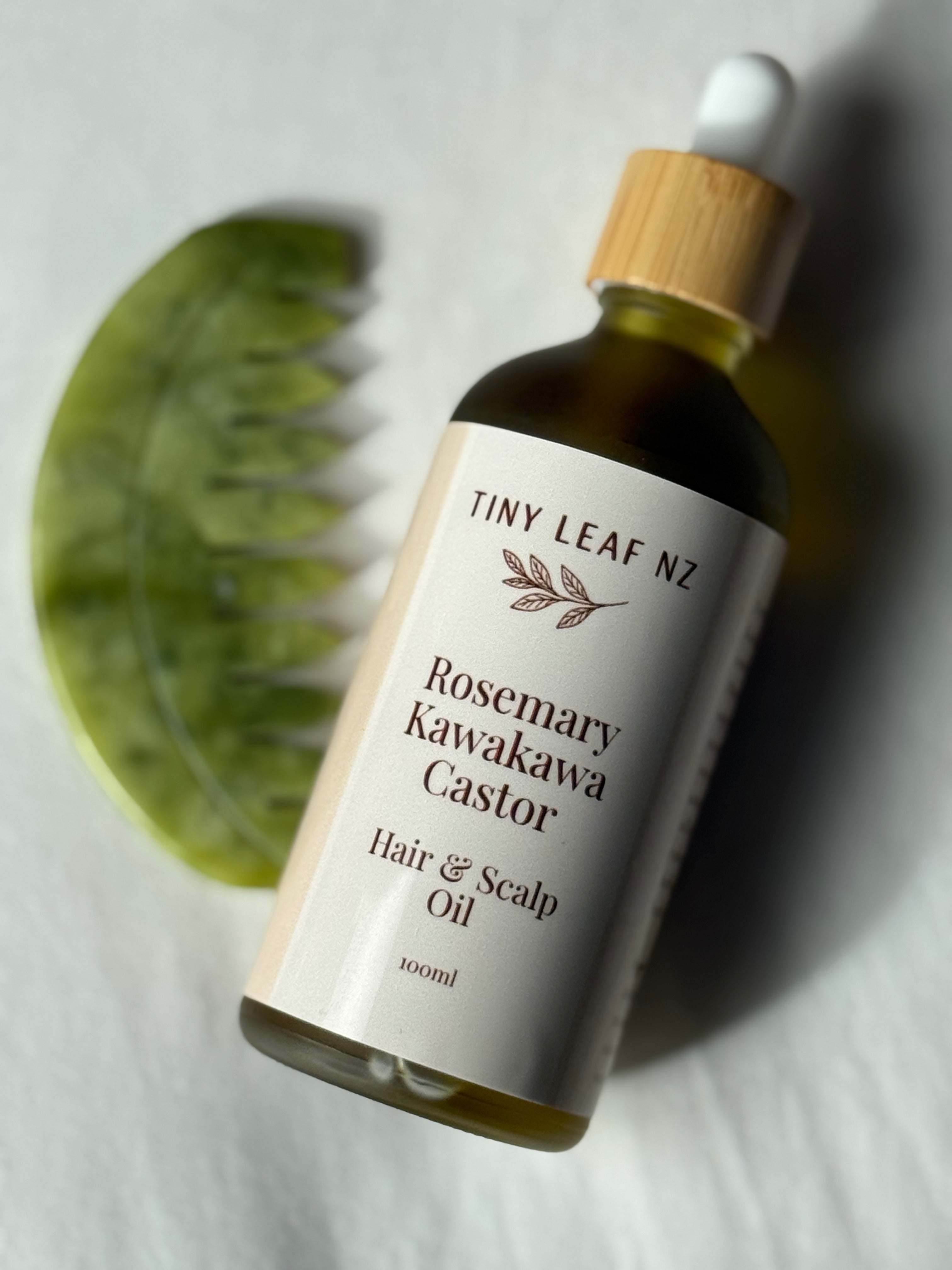 A 100ml bottle of Tiny Leaf NZ Rosemary Kawakawa Castor Hair & Scalp Oil, featuring a cream-colored label, amber glass bottle, and bamboo dropper cap, styled with a green comb on a white textured background.