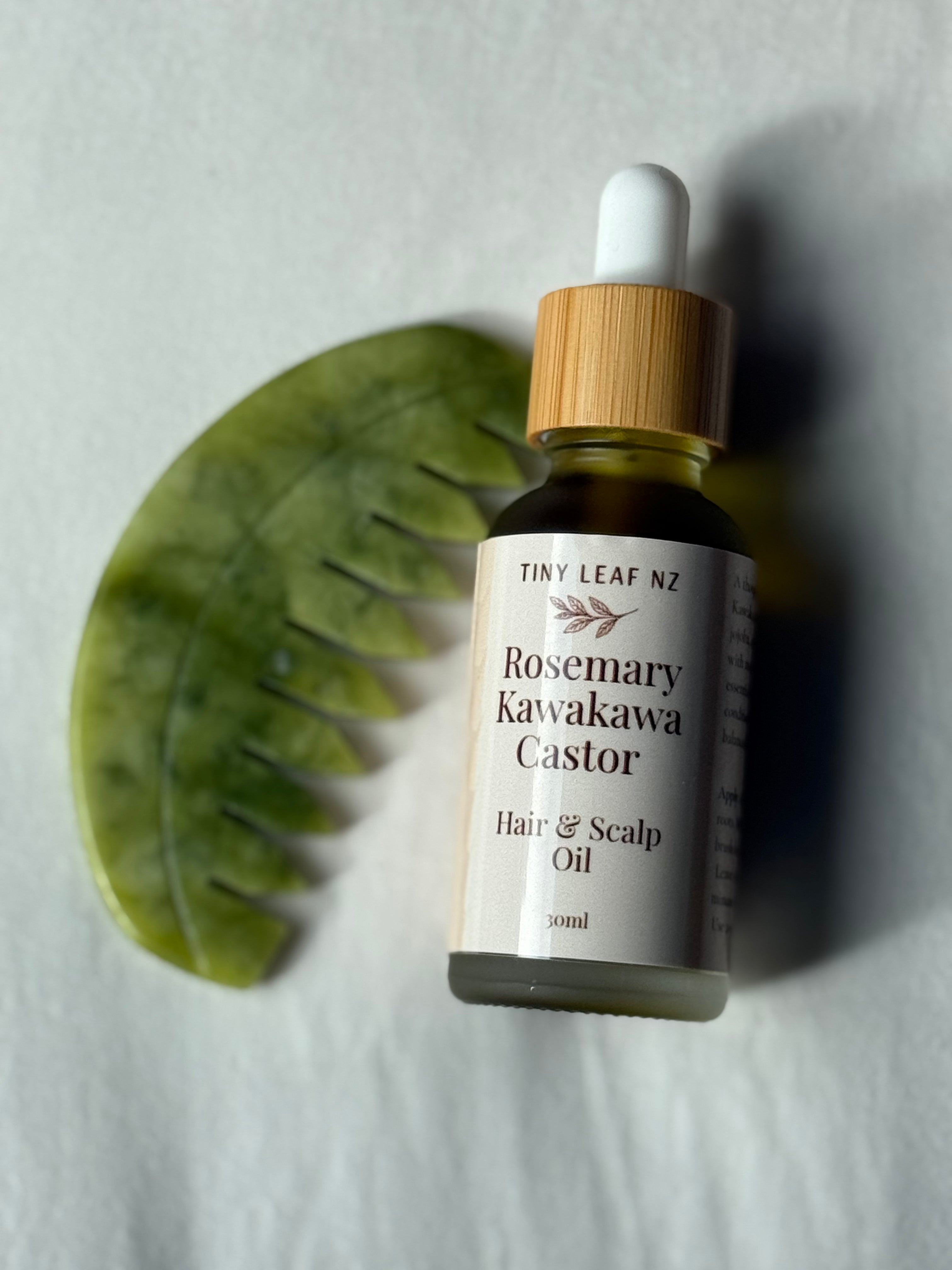 A small glass dropper bottle of Tiny Leaf NZ Rosemary Kawakawa Castor Hair & Scalp Oil, featuring a bamboo-colored dropper cap and a white label, styled next to a green leaf comb on a light gray background.