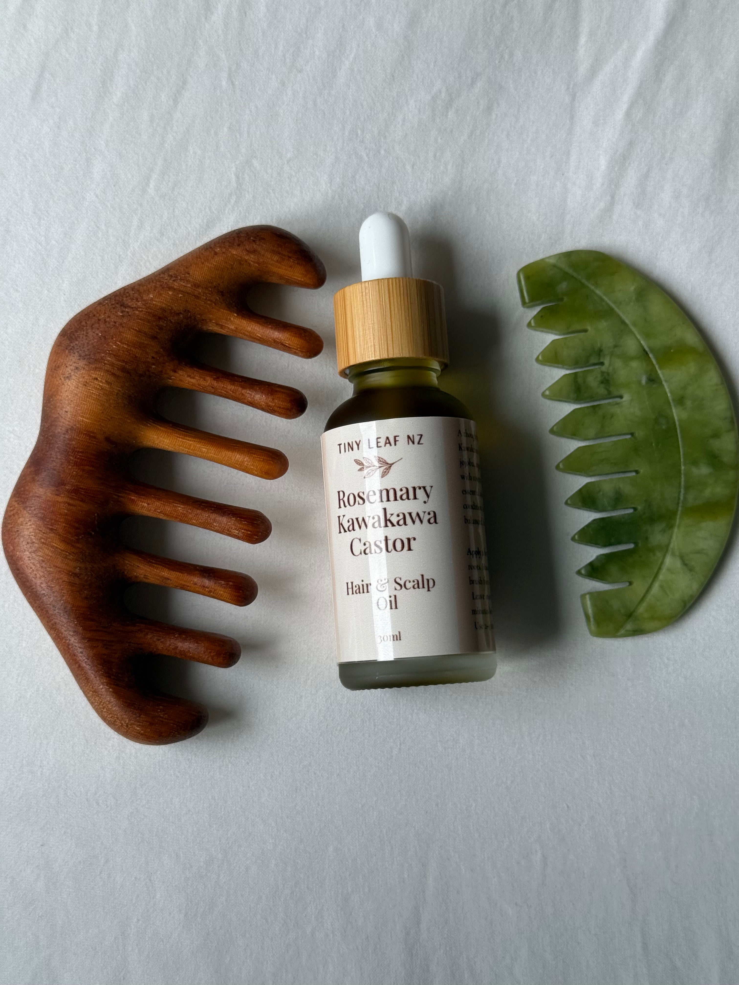 A bottle of Tiny Leaf NZ Rosemary and Kawakawa Castor Hair Scalp Oil is displayed on a light gray textured background, flanked by a wooden hair comb on the left and a green jade scalp comb on the right.
