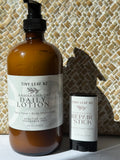 A 500 mL amber bottle of Tiny Leaf NZ Kawakawa Oat Daily Lotion stands next to a matching 60 g stick of Tiny Leaf NZ Kawakawa Repair Stick against a woven basket background.