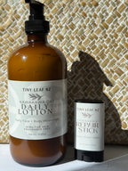A 500 mL amber bottle of Tiny Leaf NZ Kawakawa Oat Daily Lotion stands next to a matching 60 g stick of Tiny Leaf NZ Kawakawa Repair Stick against a woven basket background.