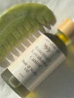 A bottle of Rosemary Kawakawa Hair & Scalp Oil by Tiny Trees, featuring a white label and a light wood cap, styled with a green leaf on a textured white surface.
