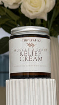 A jar of Tiny Leaf Nz Muscle & Joint Relief Cream displayed on a white ribbed pedestal with blurred white roses in the background.
