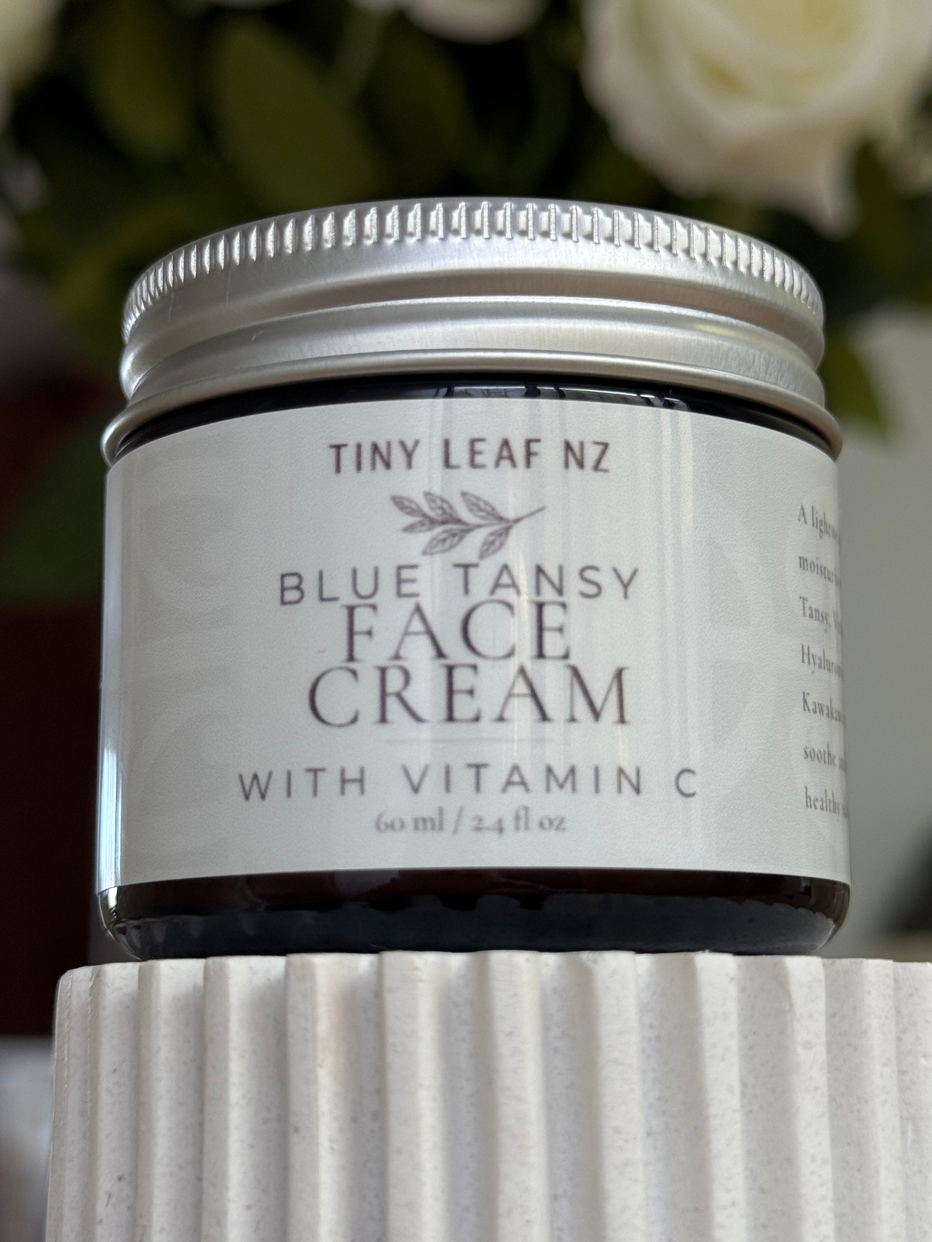 A jar of Tiny Leaf Nz Blue Tansy Face Cream with Vitamin C, featuring a white label and silver metal lid, displayed on a textured white pedestal with a blurred background of green leaves and white flowers.