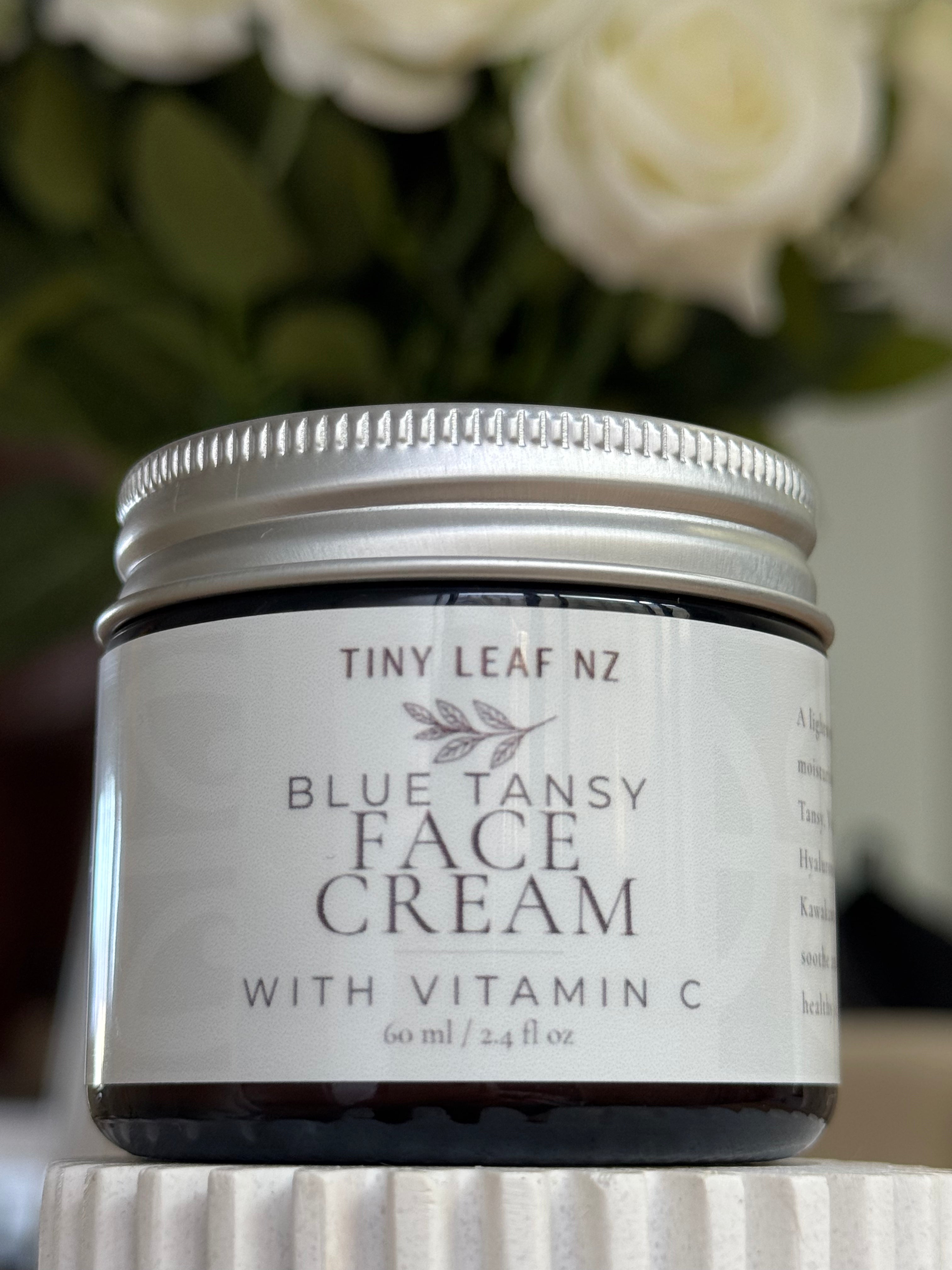 A jar of Tiny Leaf NZ Blue Tansy Face Cream with Vitamin C, featuring a white label and silver metal lid, set against a blurred background of white roses.