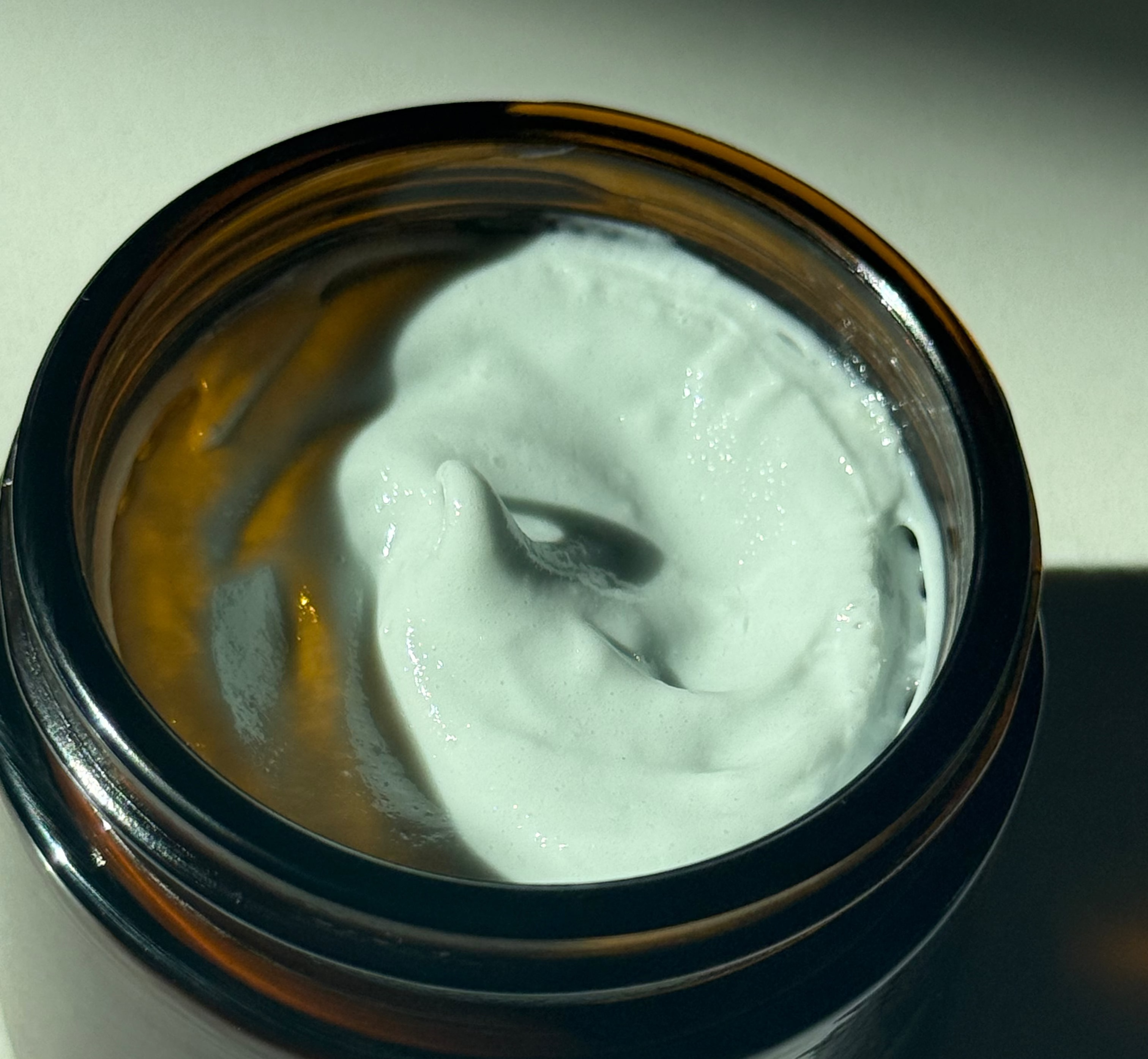 An open amber glass jar filled with thick white cream, photographed in dramatic side lighting on a light surface.