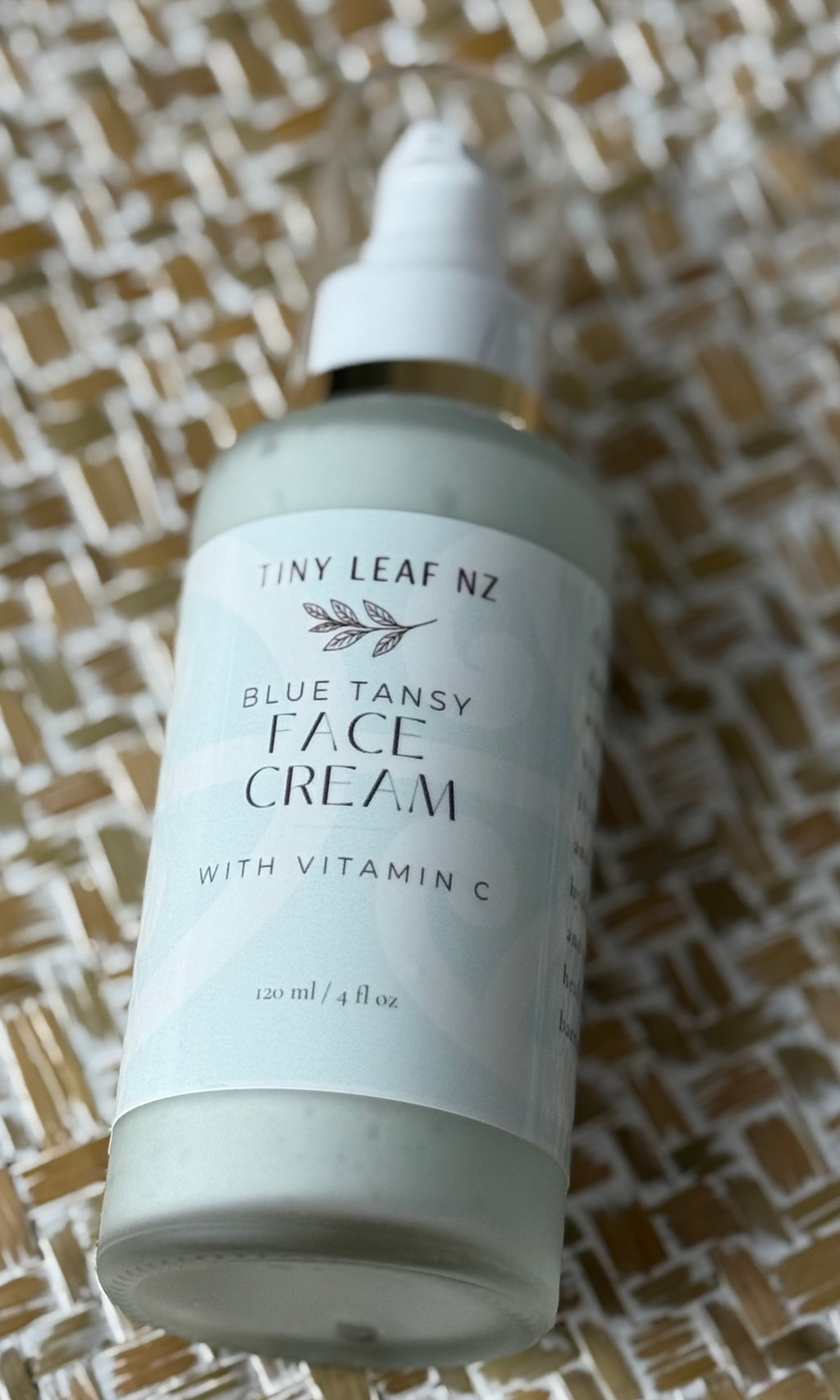 A frosted glass bottle of Tiny Leaf NZ Blue Tansy Face Cream with Vitamin C, featuring a white dropper cap and a white label, resting on a woven beige and brown mat.