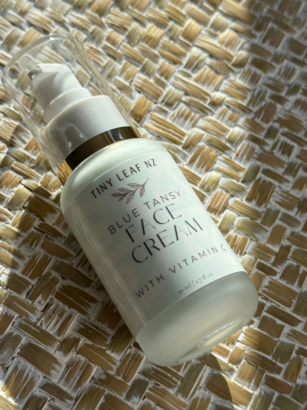 A bottle of Tiny Leaf NZ Blue Tansy Face Cream with Vitamin C, resting on a woven straw mat under warm sunlight.
