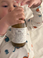 A close-up lifestyle shot of a child wearing a dinosaur-print pajama top, holding a bottle of Tiny Leaf Nz Kawakawa Oat Daily Lotion.
