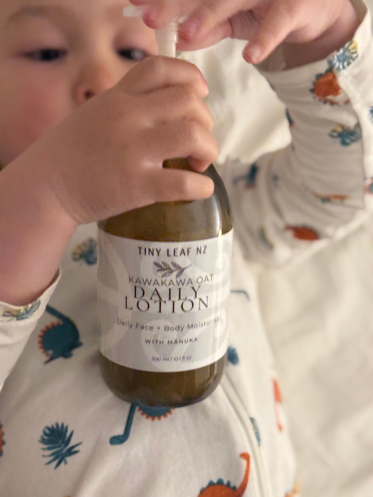 A close-up lifestyle shot of a child wearing a dinosaur-print pajama top, holding a bottle of Tiny Leaf Nz Kawakawa Oat Daily Lotion.