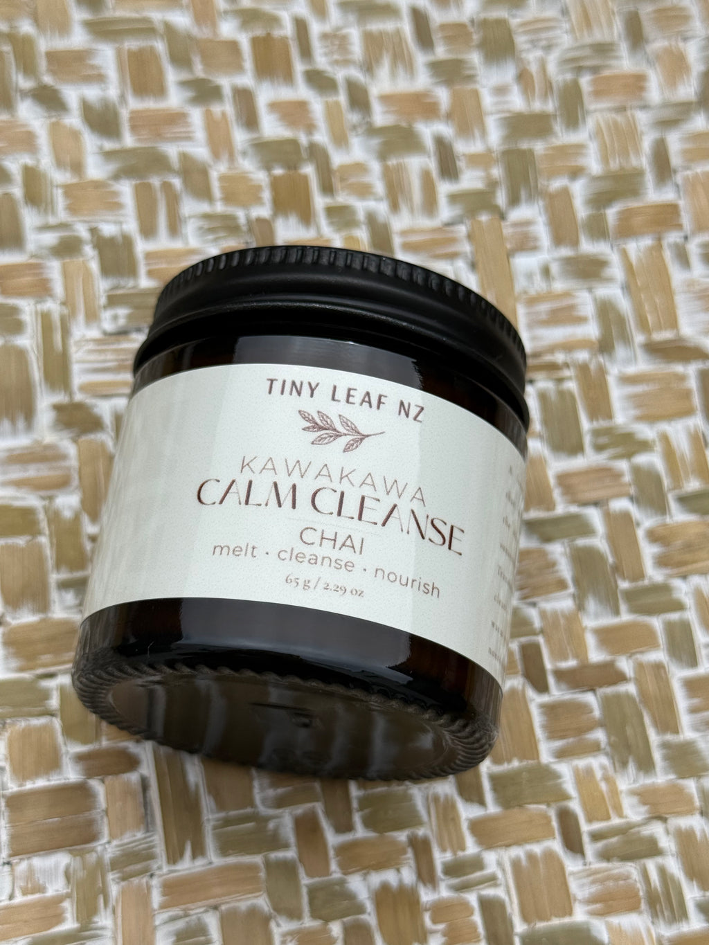 A jar of Tiny Leaf Nz Kawakawa Calm Cleanser Chai, in a dark amber glass jar with a black lid and a cream-colored label, set against a woven straw background.