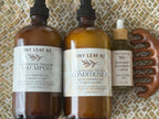 Three amber glass hair care bottles from Tiny Leaf NZ, including Shampoo, Conditioner, and Rosemary Kawaka Castor Hair of Scalp Oil, displayed alongside a wooden comb on a woven mat background.