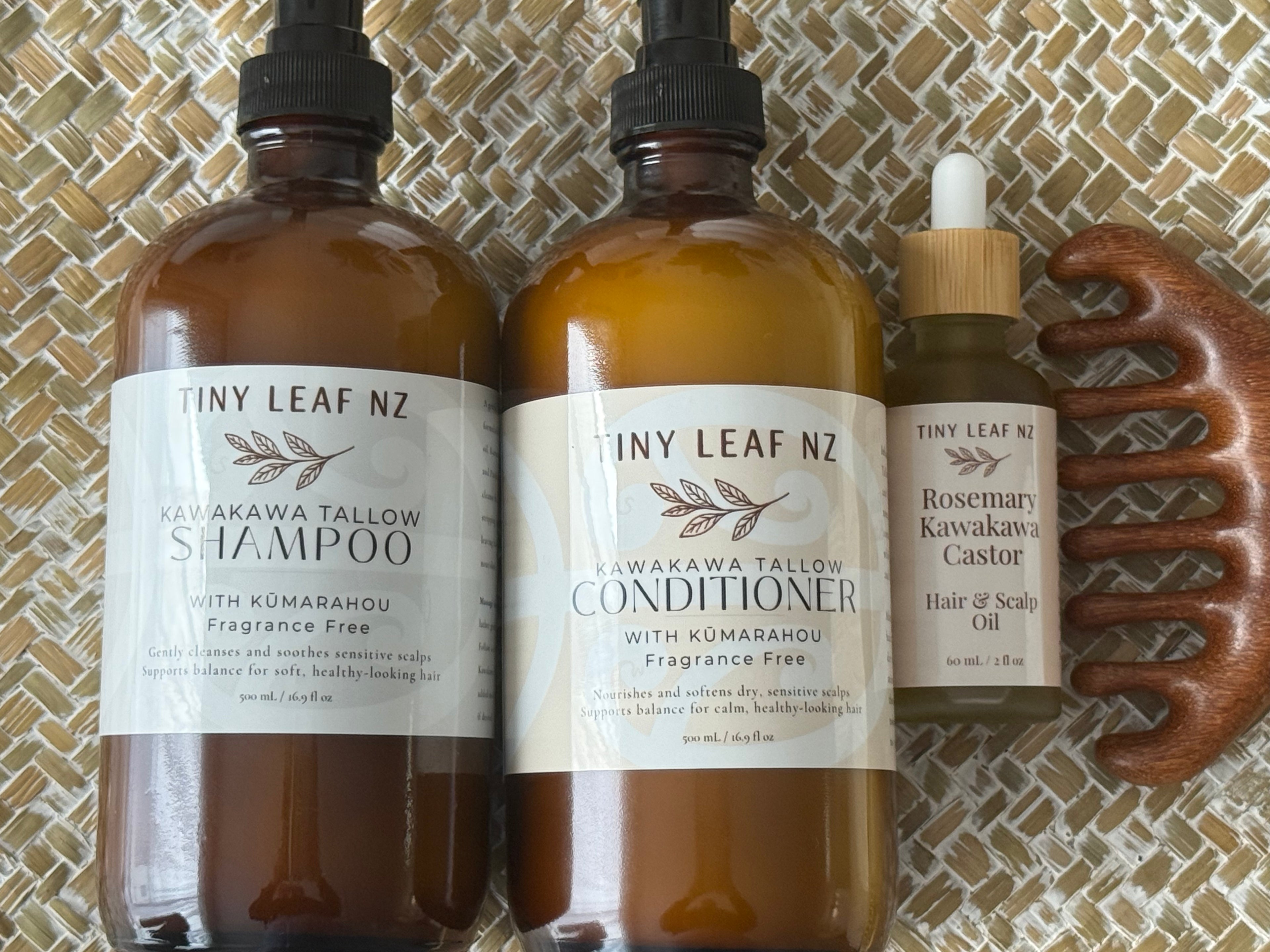 Three amber glass hair care bottles from Tiny Leaf NZ, including Shampoo, Conditioner, and Rosemary Kawaka Castor Hair of Scalp Oil, displayed alongside a wooden comb on a woven mat background.