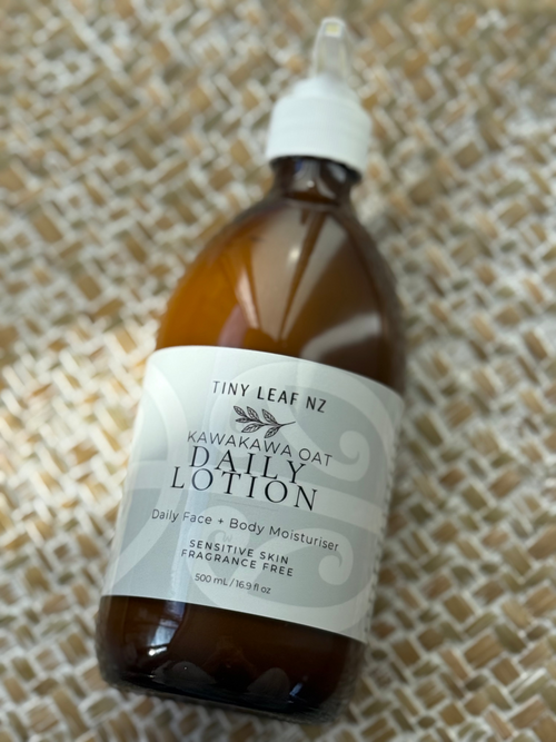 A bottle of Tiny Leaf NZ Kawakawa Oat Daily Lotion in a brown glass bottle with a white dropper cap, resting on a woven beige textured mat.