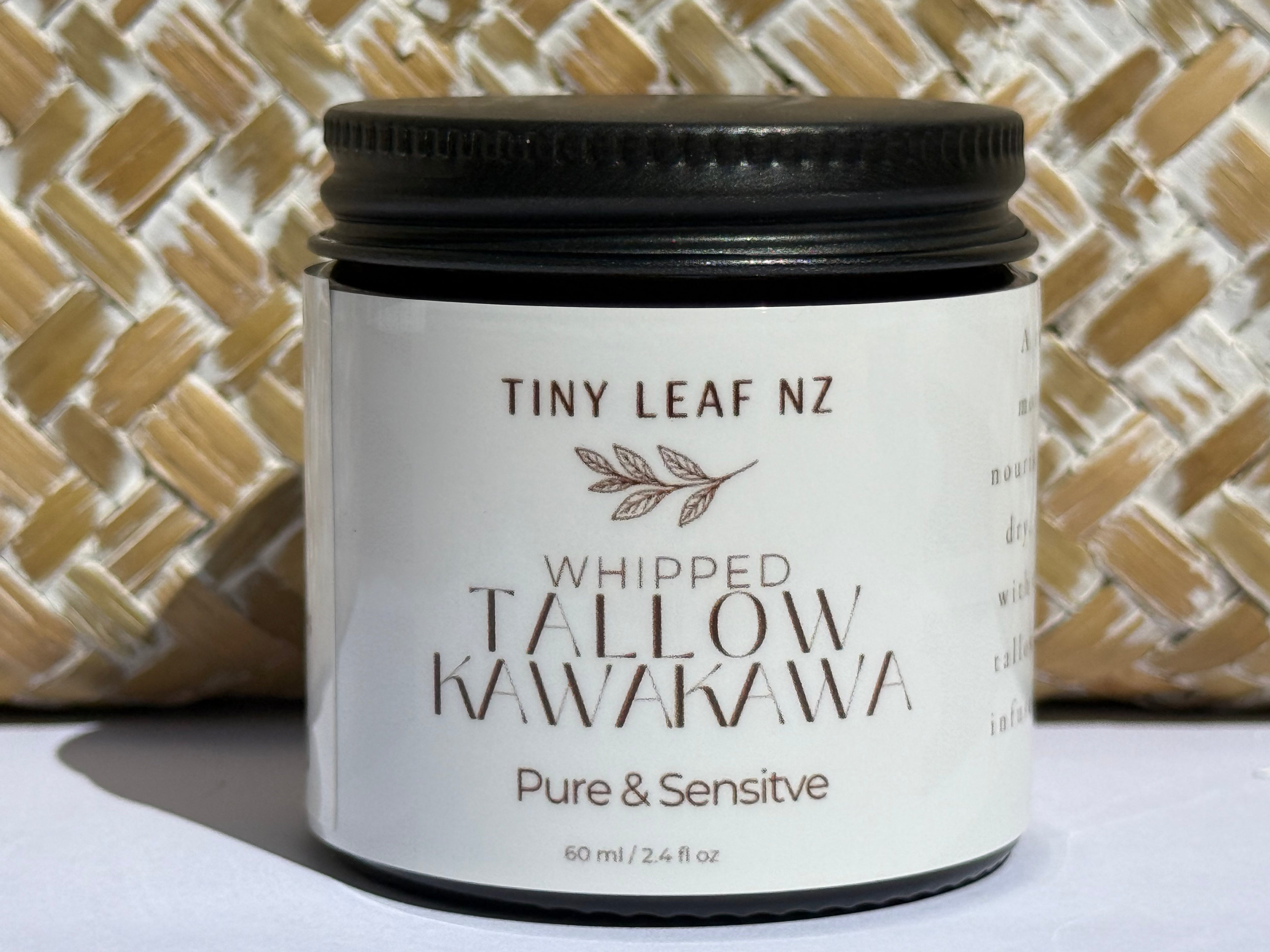 A jar of Tiny Leaf NZ Whipped Tallow Kawakawa skincare cream, featuring a white label and black lid, set against a woven straw background.
