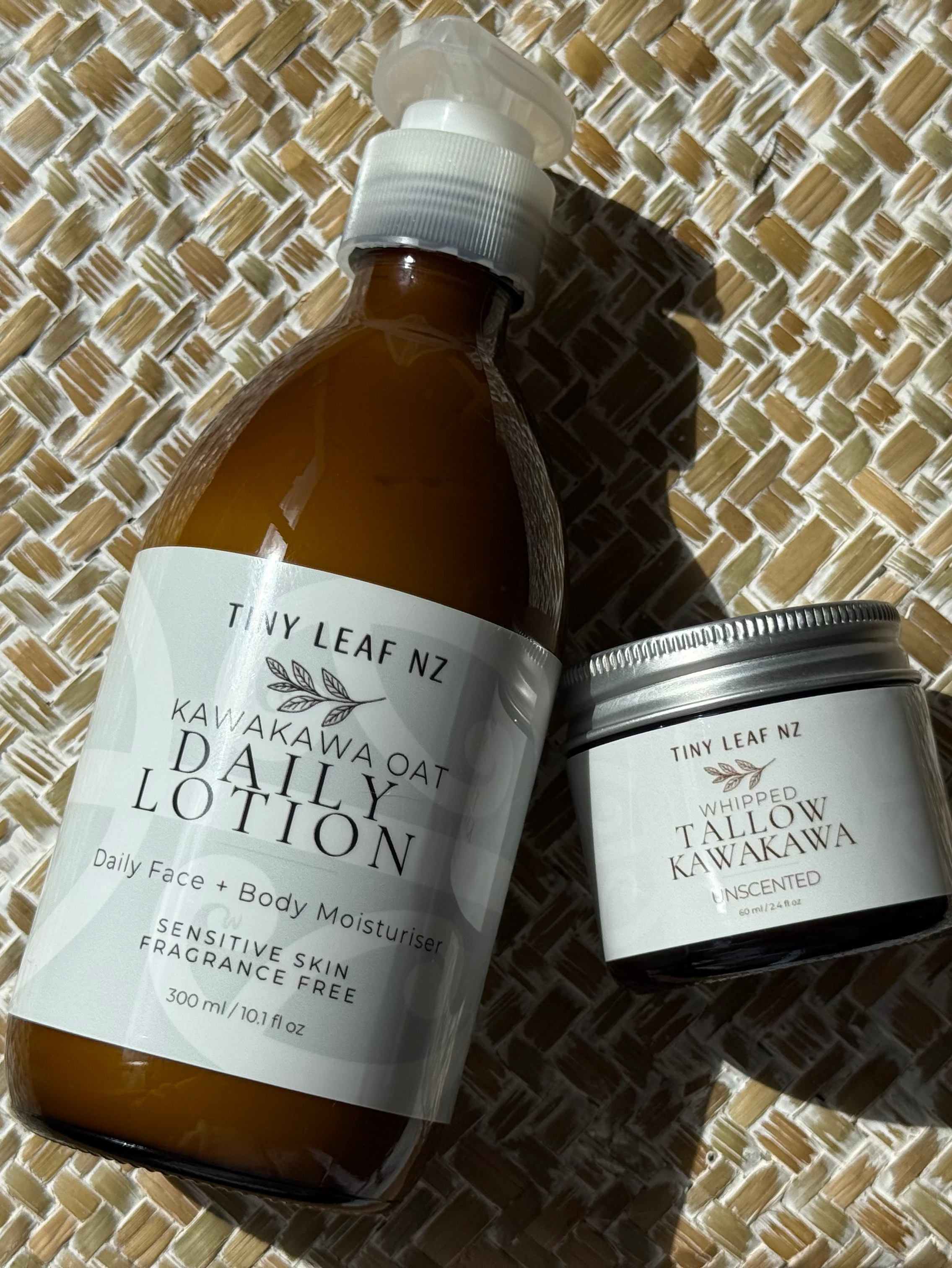 A bottle of body lotion and a small jar of body butter from the brand Tidy Leaf NZ, displayed on a woven straw mat under direct sunlight.