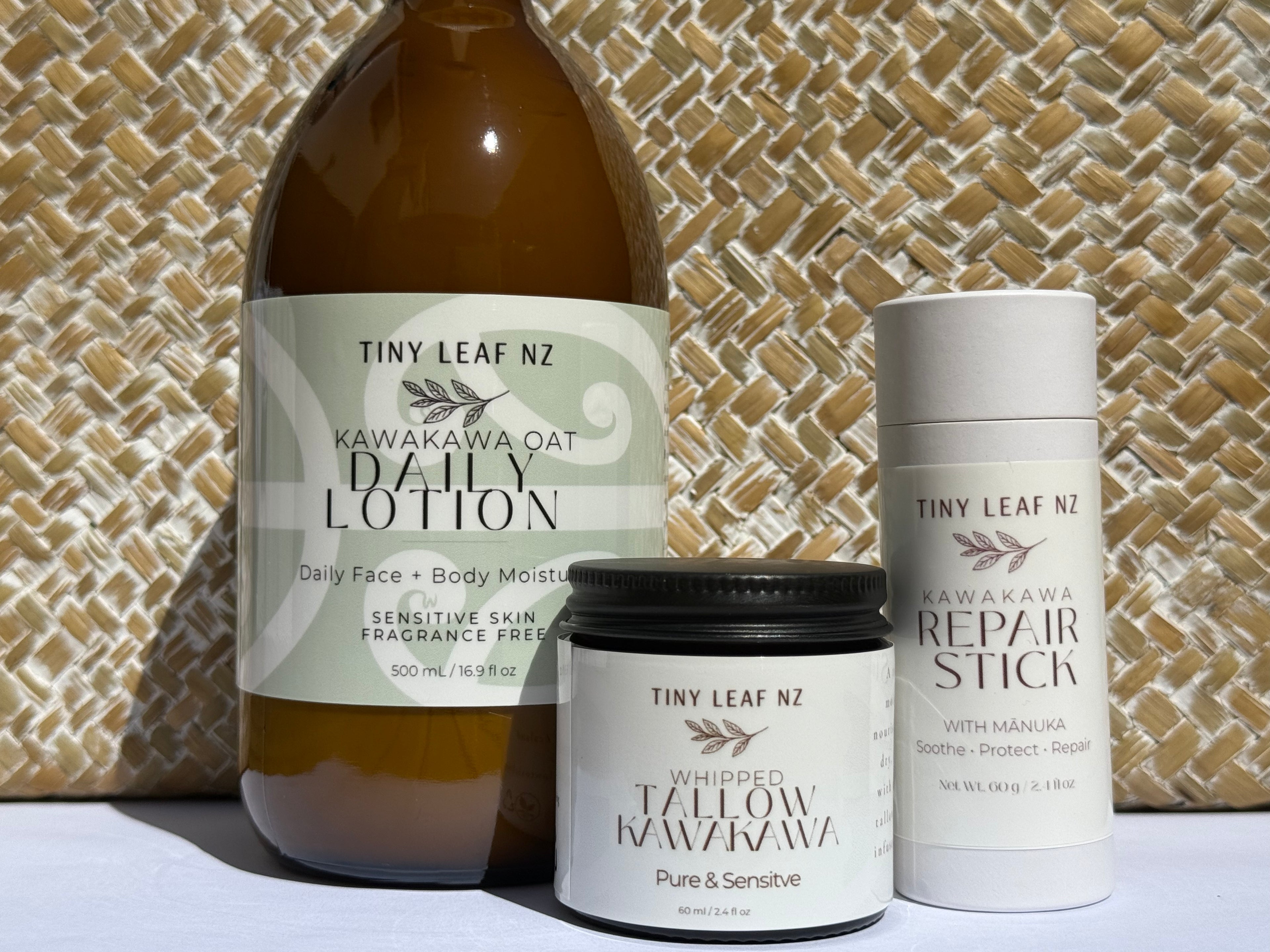 Three Tiny Leaf Nz skincare products arranged on a white surface against a woven straw background: a large amber bottle of Kawakawa Oat Daily Lotion, a white tube of Kawakawa Repair Stick, and a white jar of Whipped Tallow Kawakawa.