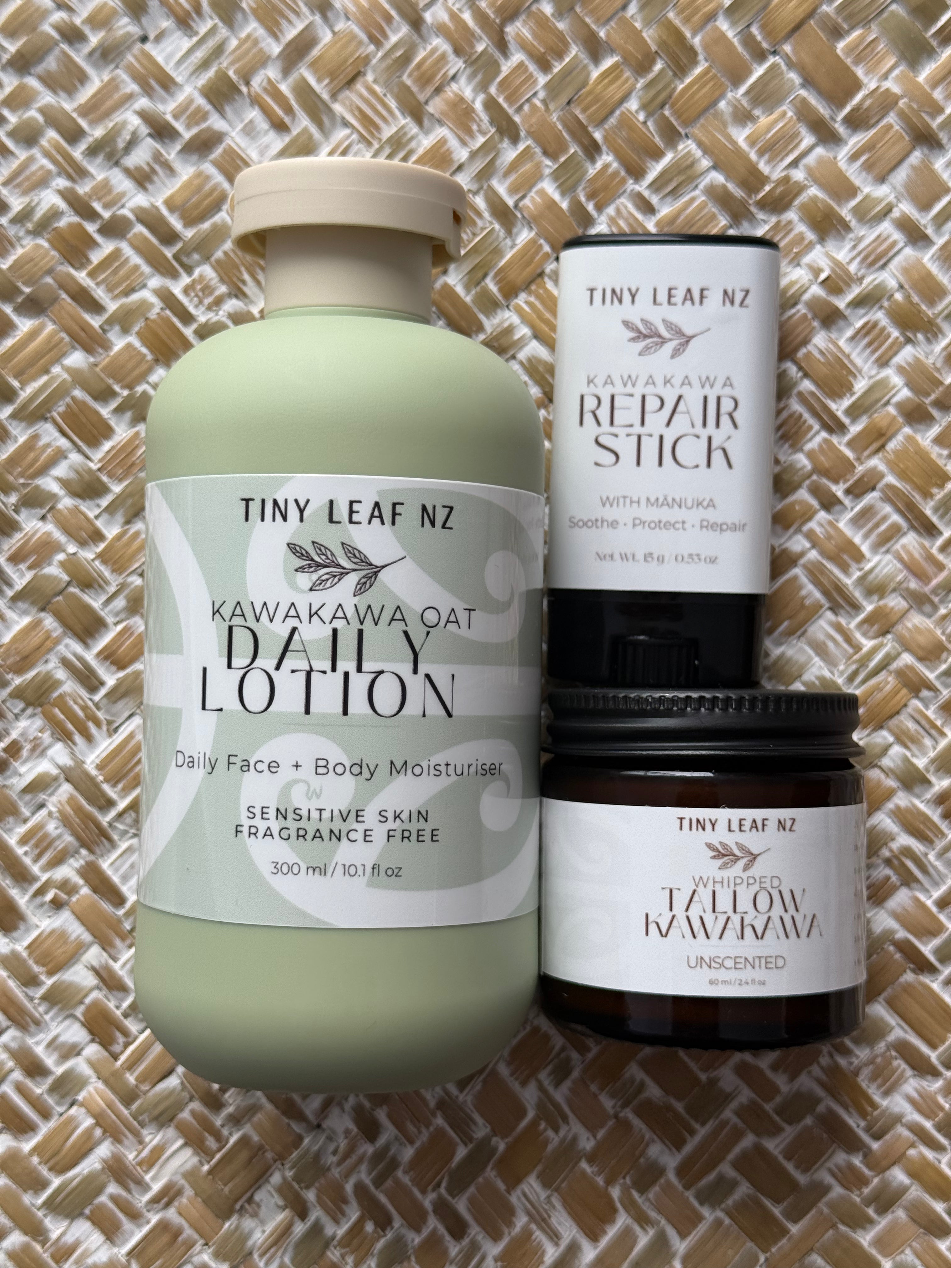 Three Tiny Leaf NZ natural skincare products arranged on a woven straw mat: a large pale green bottle of Kawakawa Oat Daily Lotion, a white tube of Kawakawa Repair Stick, and a dark amber jar of Yellow Kawakawa Unscented.
