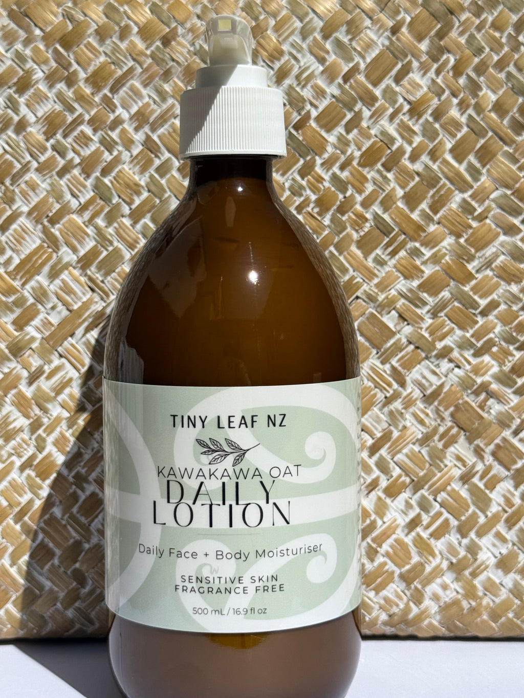 A 500ml amber glass bottle of Tiny Leaf NZ Kawakawa Oat Daily Lotion with a white spray cap, set against a woven straw background.