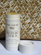 A white cylindrical skincare container labeled 'TINY LEAF NZ KAWAKAWA REPAIR STICK' stands next to its open white cap on a white surface, with a woven tan and white basket or mat behind it.