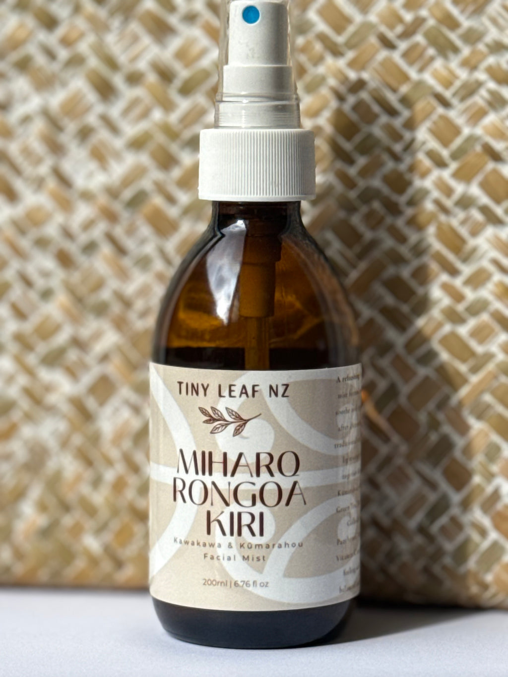 A 200ml amber glass spray bottle of Tiny Leaf Nz Miharo Rongoa Kiri facial mist, featuring a white spray nozzle and a beige label, set against a textured beige woven background.