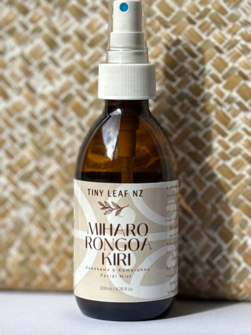 A 200ml amber glass spray bottle of Tiny Leaf Nz Miharo Rongoa Kiri facial mist, featuring a white spray nozzle and a beige label, set against a textured beige woven background.