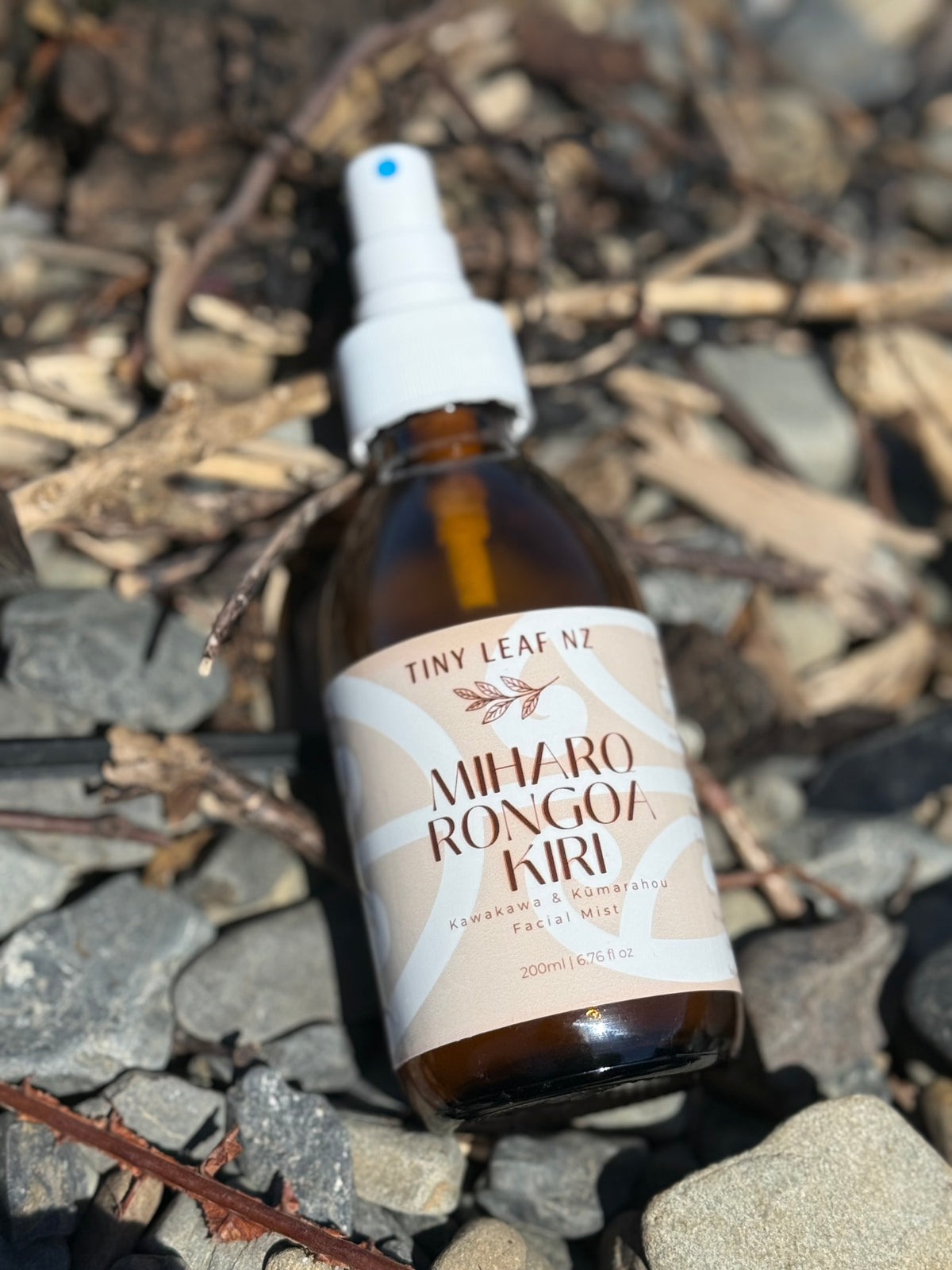 A small amber glass spray bottle with a white pump top and a cream label reading 'Tiny Leaf NZ' and 'Miharo Rongoa Kiri', placed on a bed of gray rocks and dried leaves outdoors.