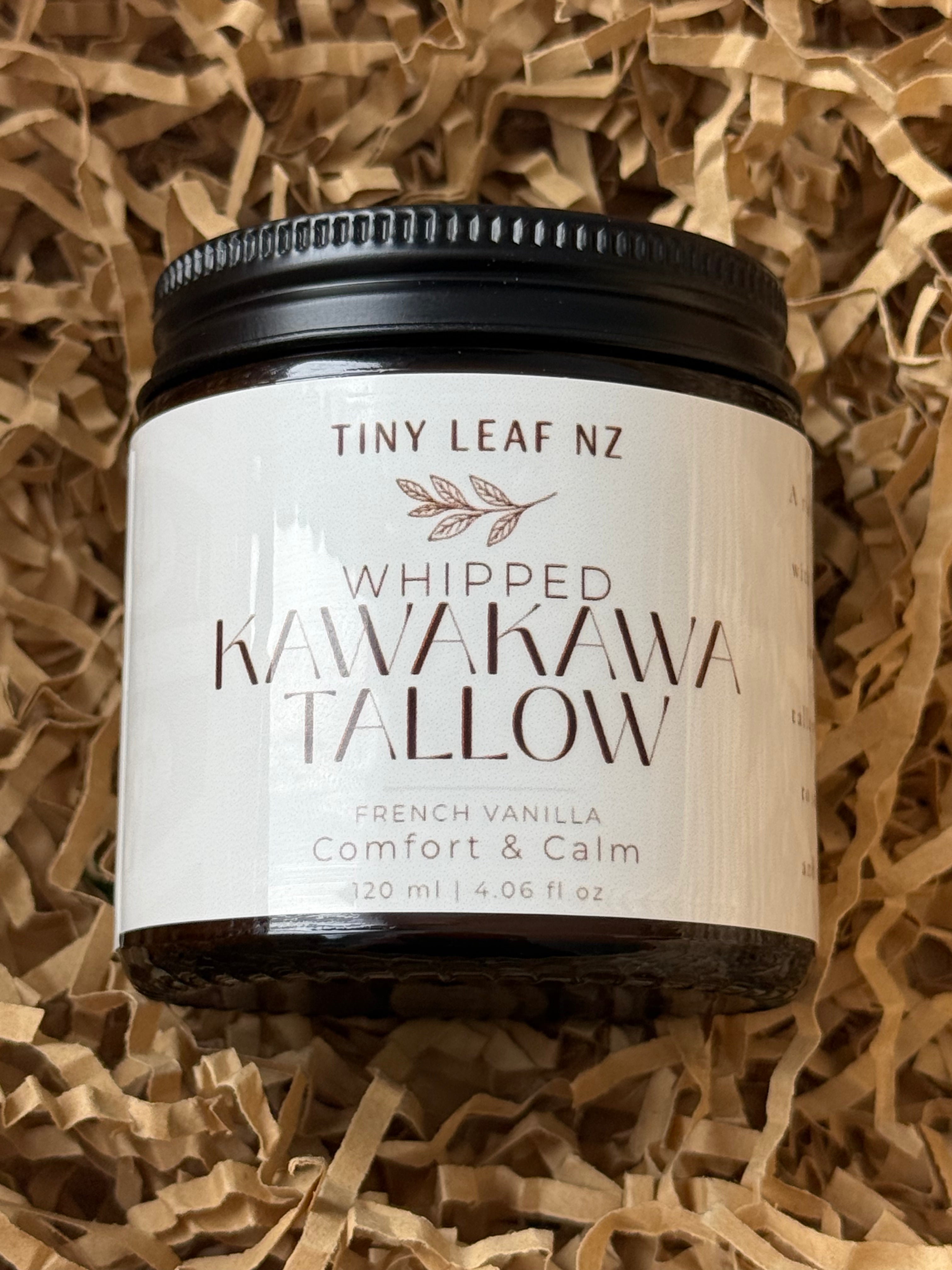 A jar of Tiny Leaf Nz Whipped Kawakawa Tallow body butter, French Vanilla, resting on a bed of brown paper shred. The product features a black lid and a cream-colored label with brown text.