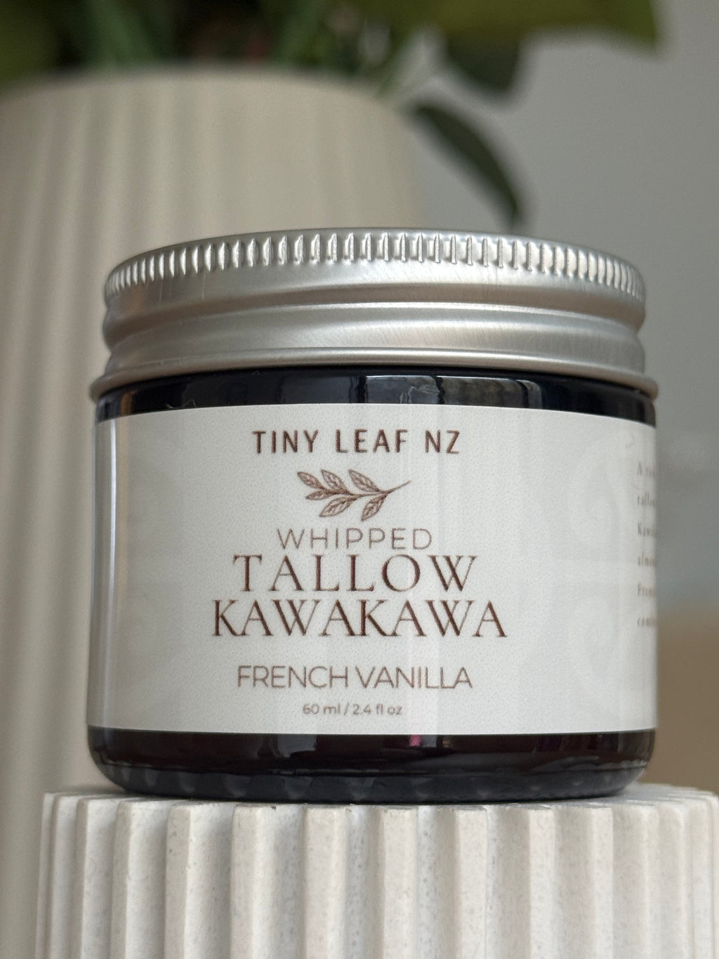 A jar of Tiny Leaf NZ Whipped Tallow Kawakawa French Vanilla balm, featuring a white label and silver metal lid, displayed on a white ribbed pedestal.