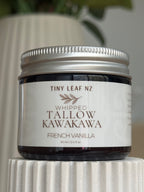 A jar of Tiny Leaf NZ Whipped Tallow Kawakawa French Vanilla balm, featuring a white label and silver metal lid, displayed on a white ribbed pedestal.
