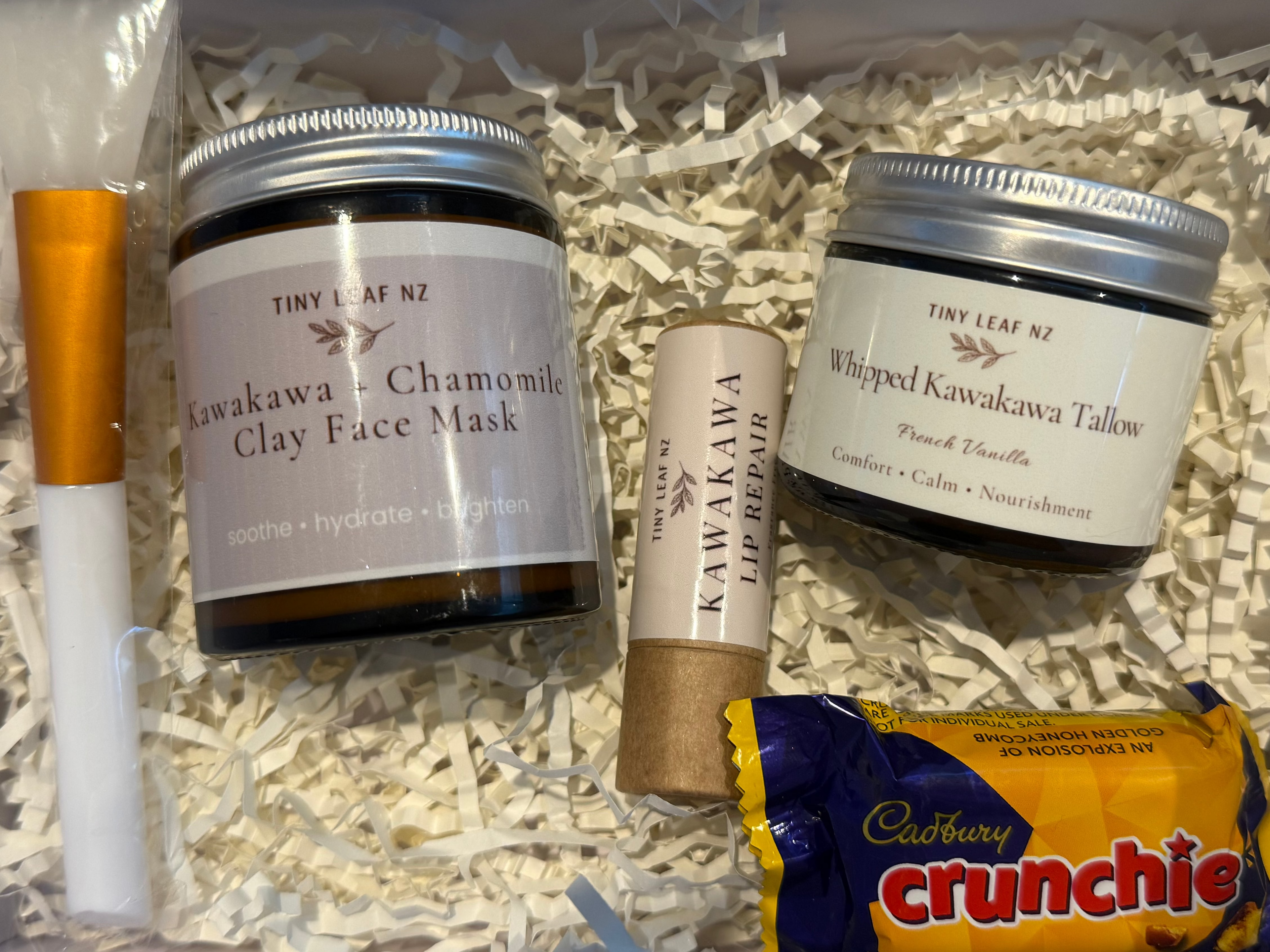 Cosmetic products including jars, a brush, and a packet of Crunchie chocolate in a box with shredded paper.