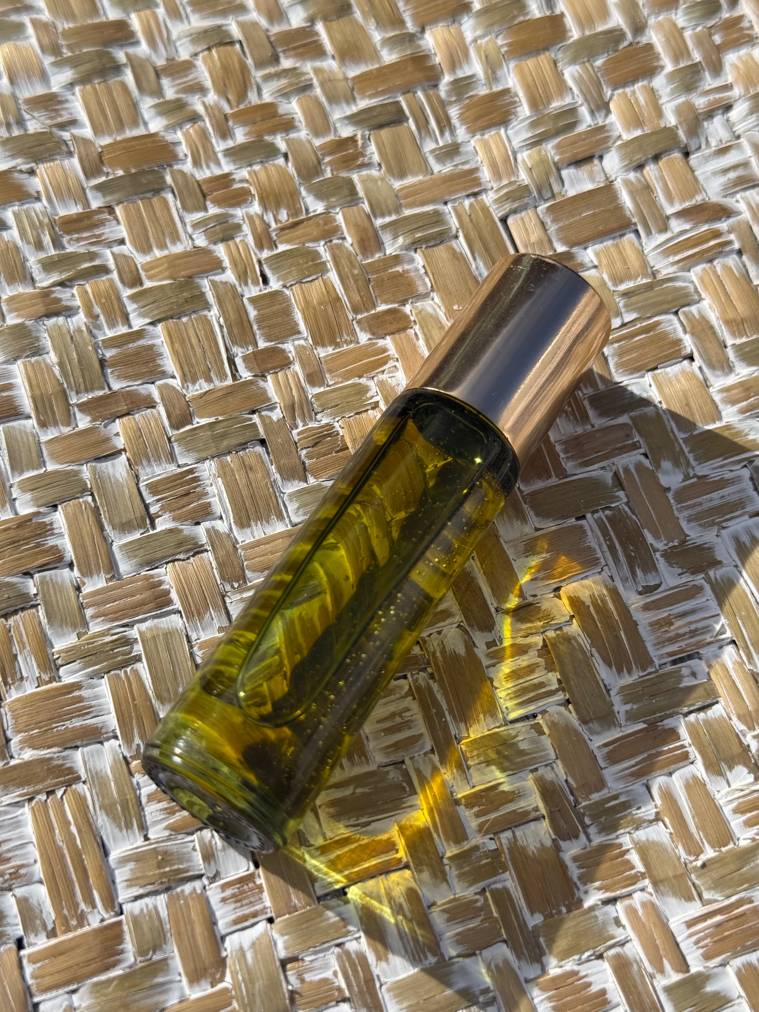 A clear glass roller bottle filled with golden-yellow liquid, topped with a reflective silver cap, resting diagonally on a woven straw mat under bright sunlight.