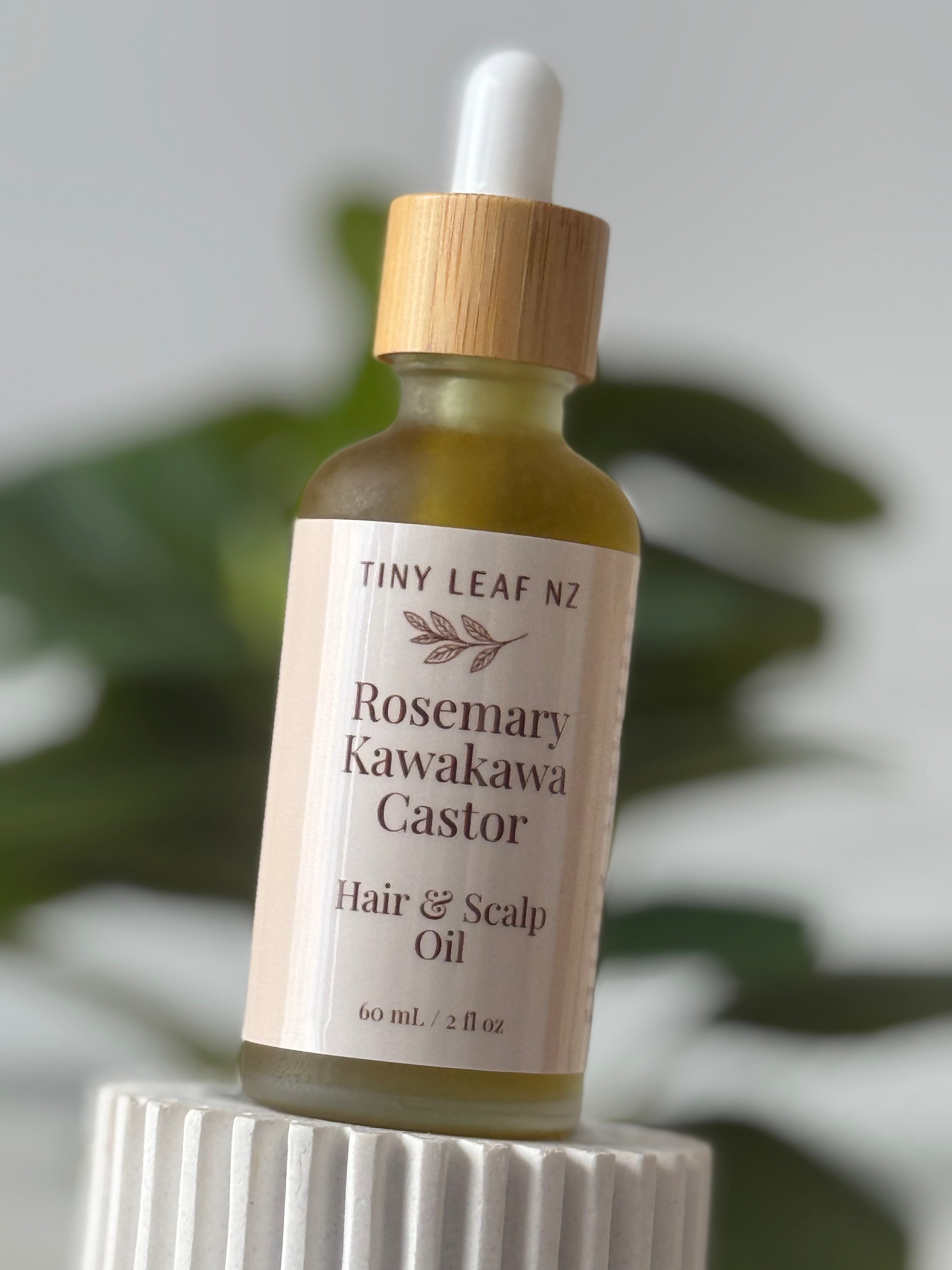A bottle of Tiny Leaf NZ Rosemary Kawakawa Castor Hair & Scalp Oil, featuring a clear glass bottle with a light wood dropper cap and a cream-colored label, displayed on a white ribbed pedestal with a blurred green plant in the background.