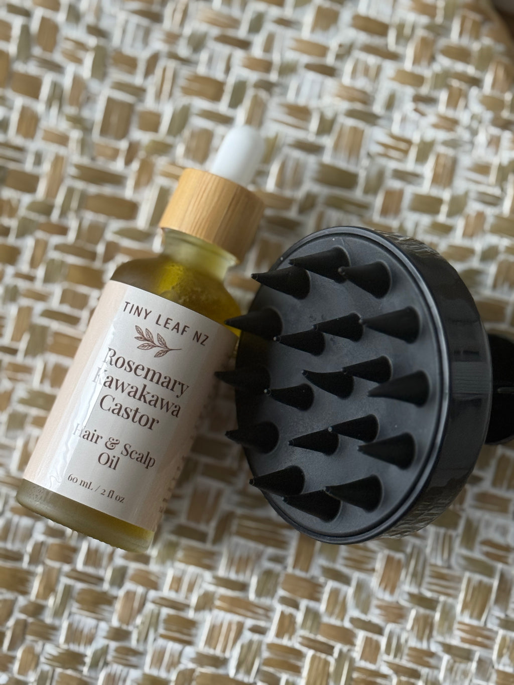 Tiny Leaf Nz Rosemary Awakawa Castor Hair & Scalp Oil bottle with a bamboo dropper cap next to a black silicone scalp massager brush, set on a woven straw mat background.