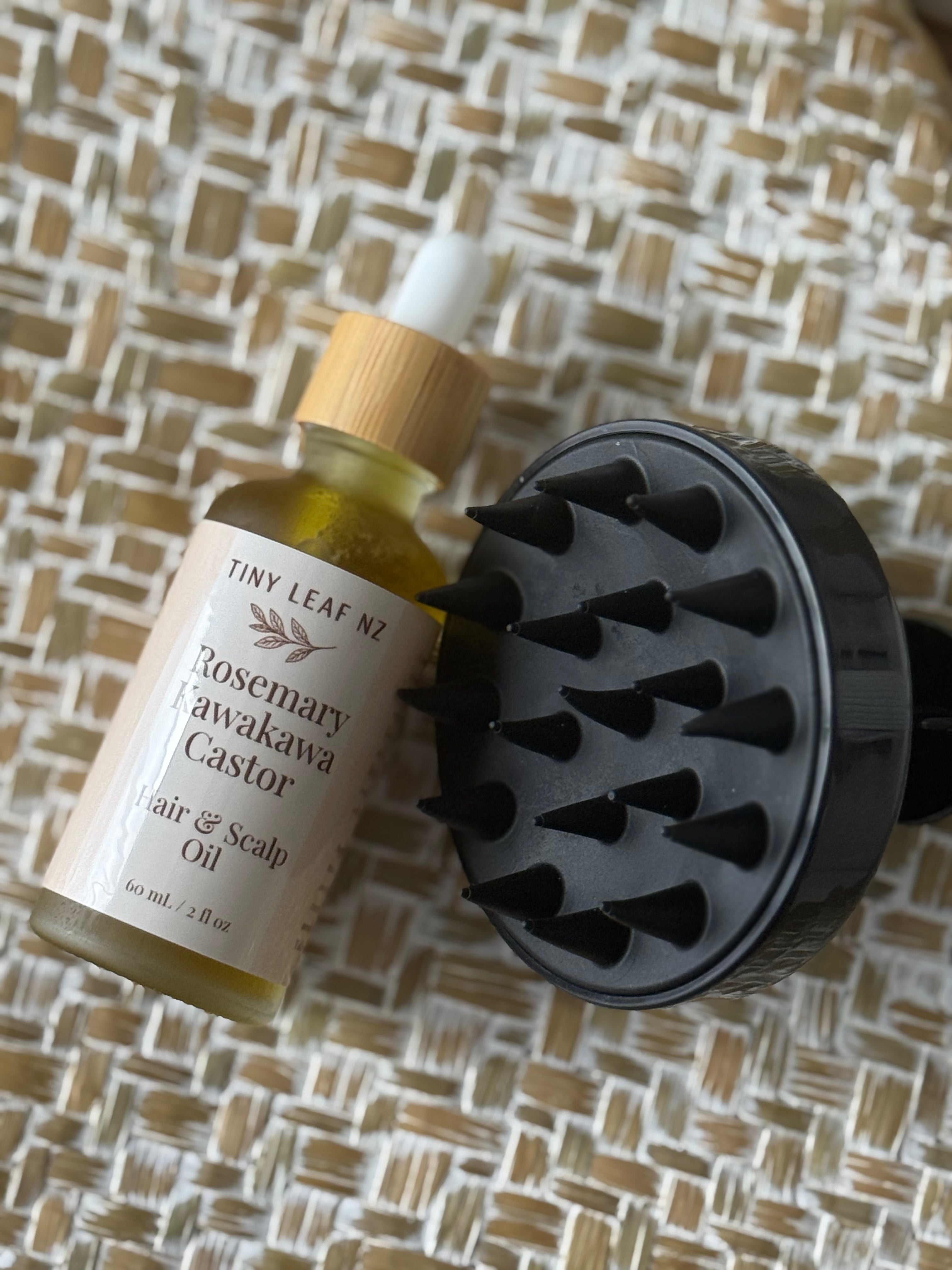 Tiny Leaf Nz Rosemary Awakawa Castor Hair & Scalp Oil bottle with a bamboo dropper cap next to a black silicone scalp massager brush, set on a woven straw mat background.