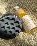 A bottle of Tiny Leaf NZ Rosemary Kawakawa Hair & Scalp Oil next to a black silicone scalp massager, arranged on a woven straw mat background.