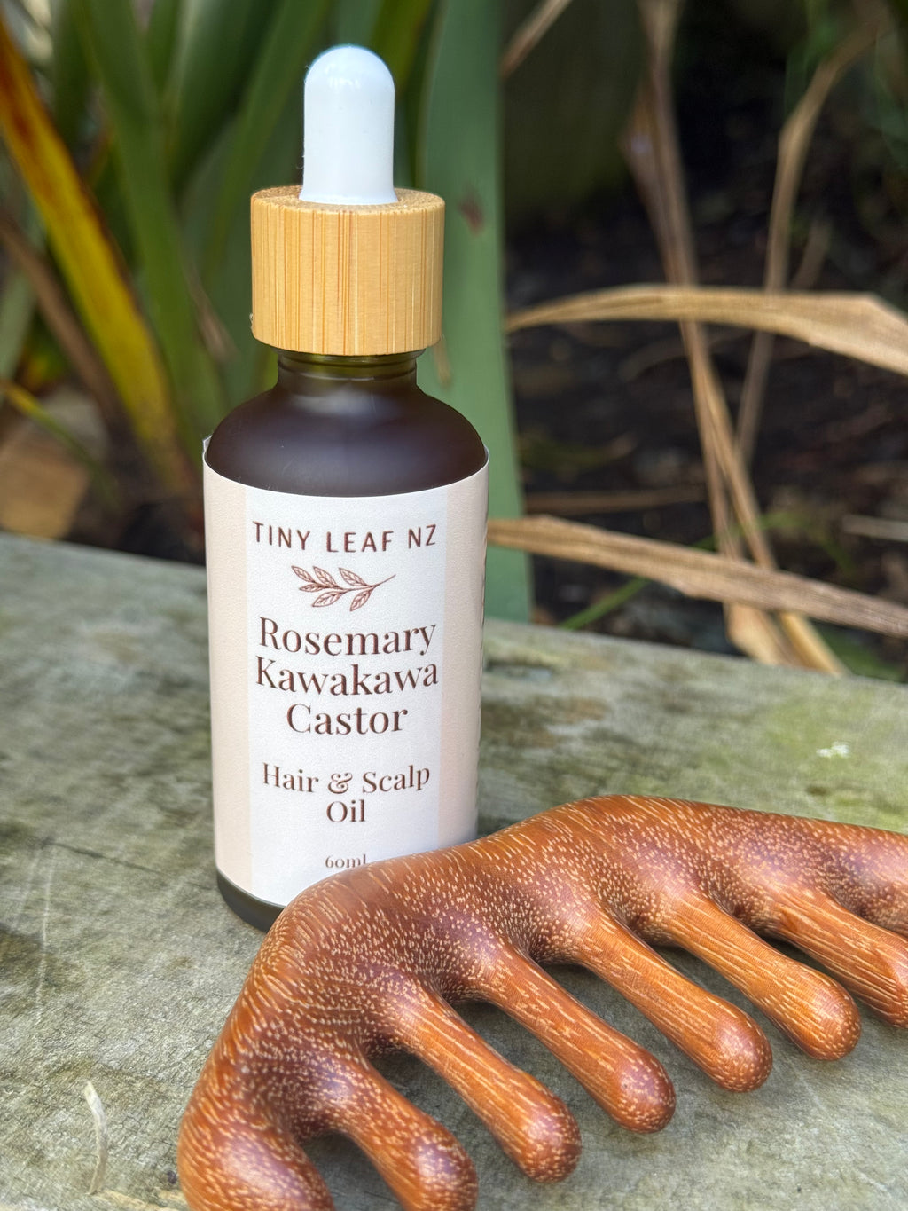 A bottle of Tiny Leaf NZ Rosemary Kawakawa Castor Hair & Scalp Oil stands on a stone surface outdoors, with a wooden scalp comb placed in front. The background shows blurred greenery.