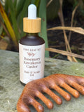 A bottle of Tiny Leaf NZ Rosemary Kawakawa Castor Hair & Scalp Oil stands on a stone surface outdoors, with a wooden scalp comb placed in front. The background shows blurred greenery.
