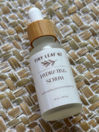 A 30 mL dropper bottle of Tiny Leaf Nz Hydrating Serum, featuring a white label and a light bamboo-colored wooden dropper cap, resting on a woven straw mat background.