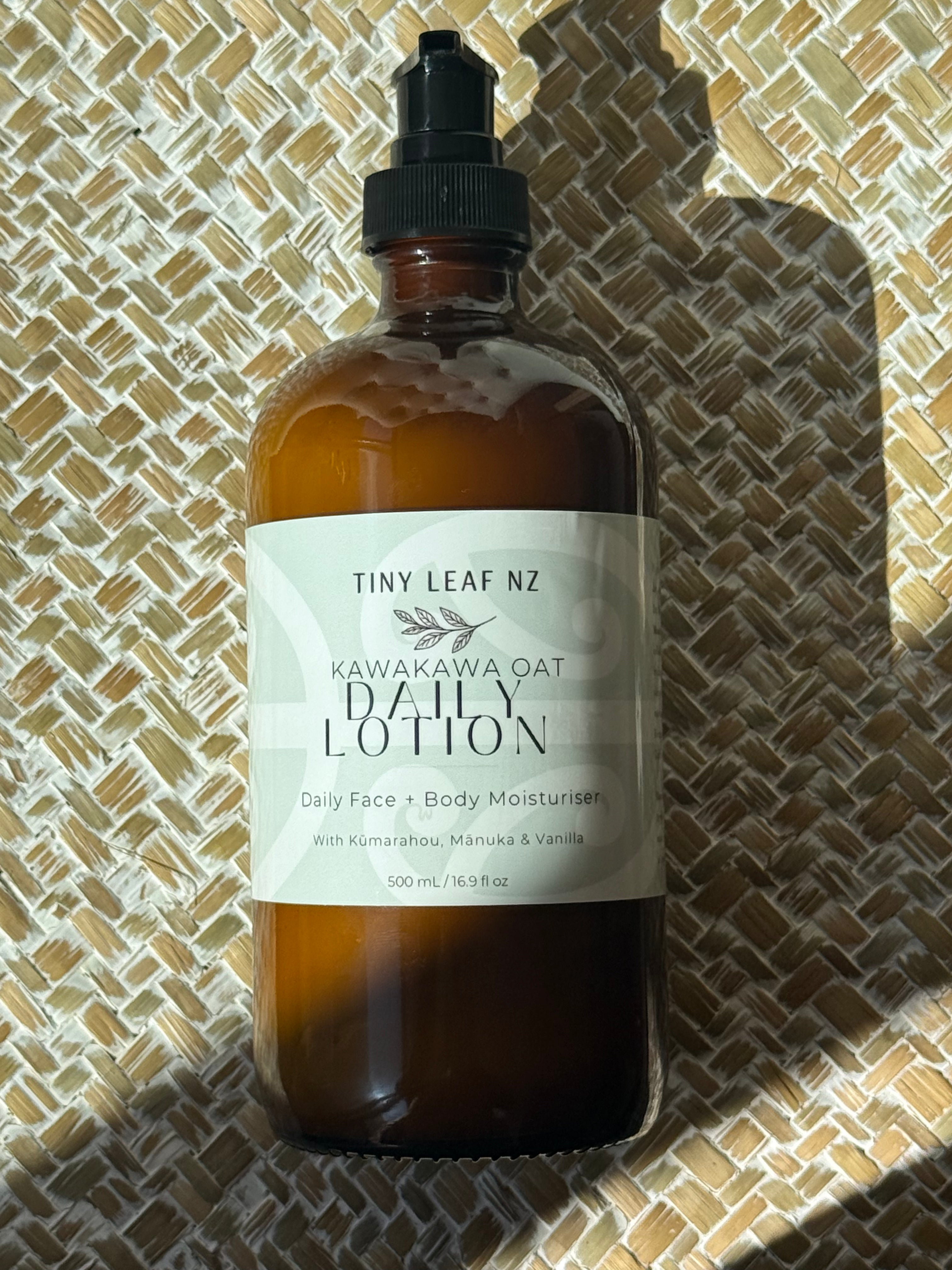 A 500ml amber glass bottle of Tiny Leaf NZ Kawakawa Oat Daily Lotion with a black dropper cap, resting on a woven straw mat under direct sunlight.