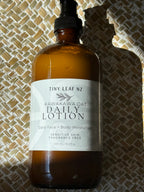 A 500ml amber glass bottle of Tiny Leaf NZ Kawakawa Oat Daily Lotion with a black spray cap, resting on a textured woven mat under direct sunlight.