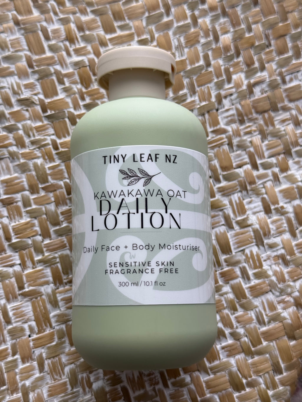 A bottle of Tiny Leaf Nz Kawakawa Oat Daily Lotion in a pale sage green color with a white label, placed on a woven straw mat background.