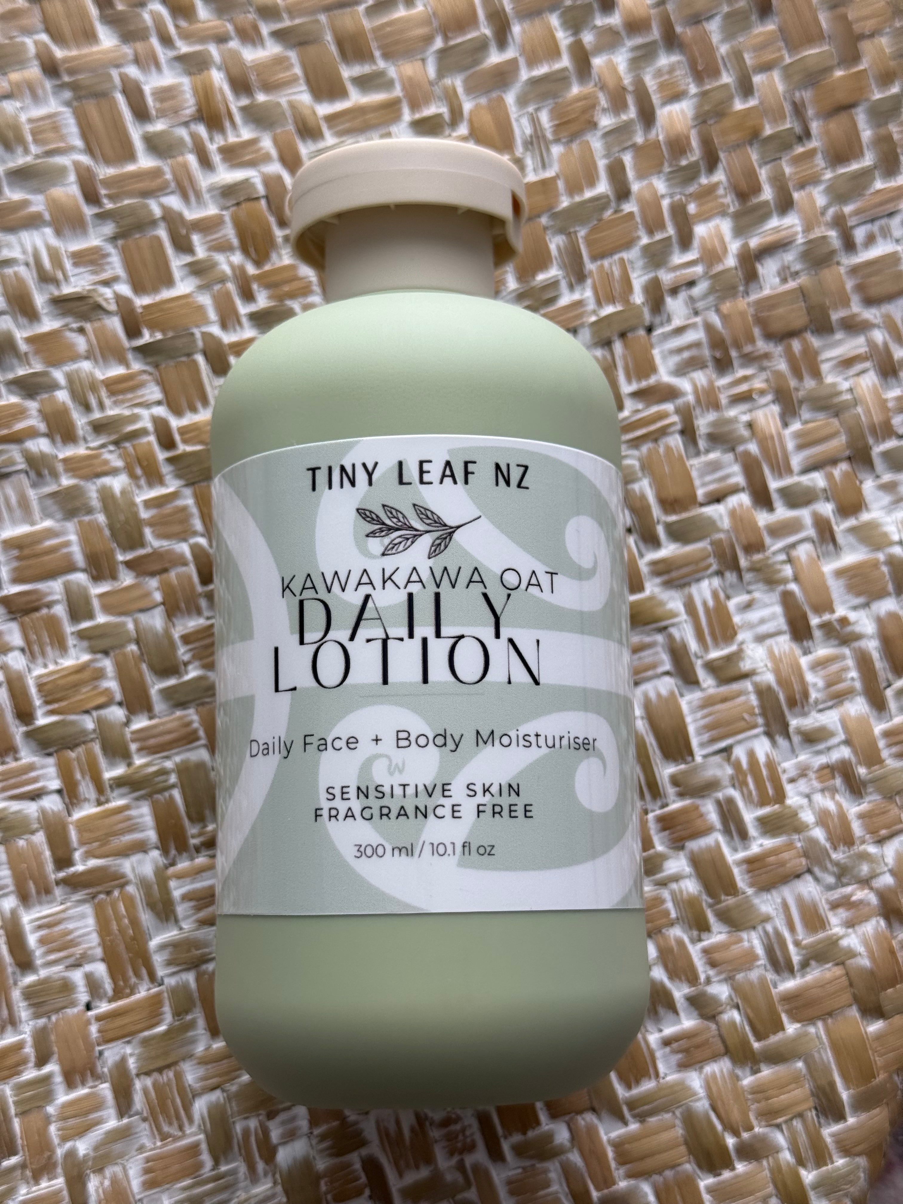 A bottle of Tiny Leaf Nz Kawakawa Oat Daily Lotion in a pale sage green color with a white label, placed on a woven straw mat background.