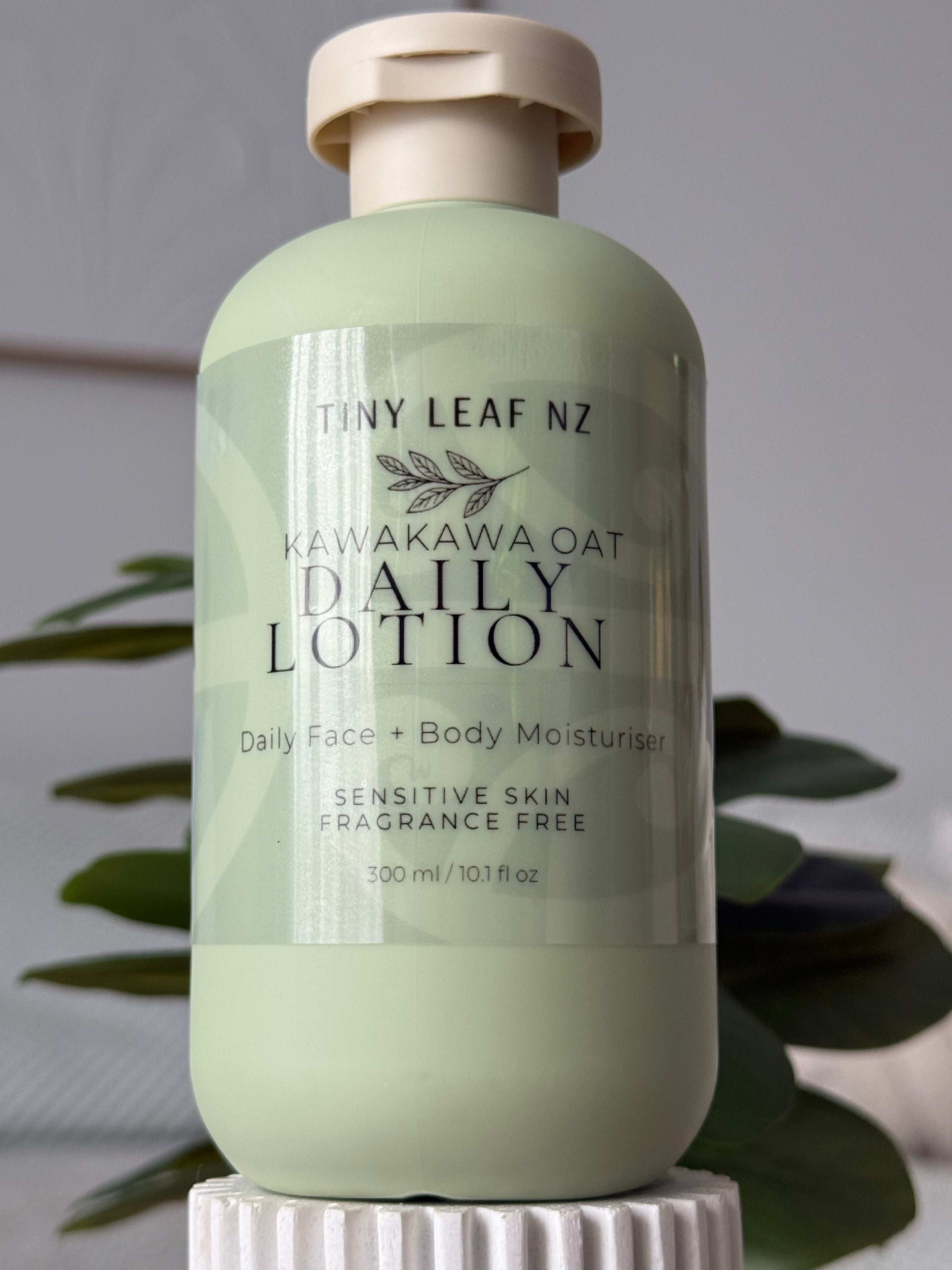 A bottle of Tiny Leaf Nz Kawakawa Oat Daily Lotion in a pale sage green color, displayed on a white pedestal with blurred green leaves in the background.
