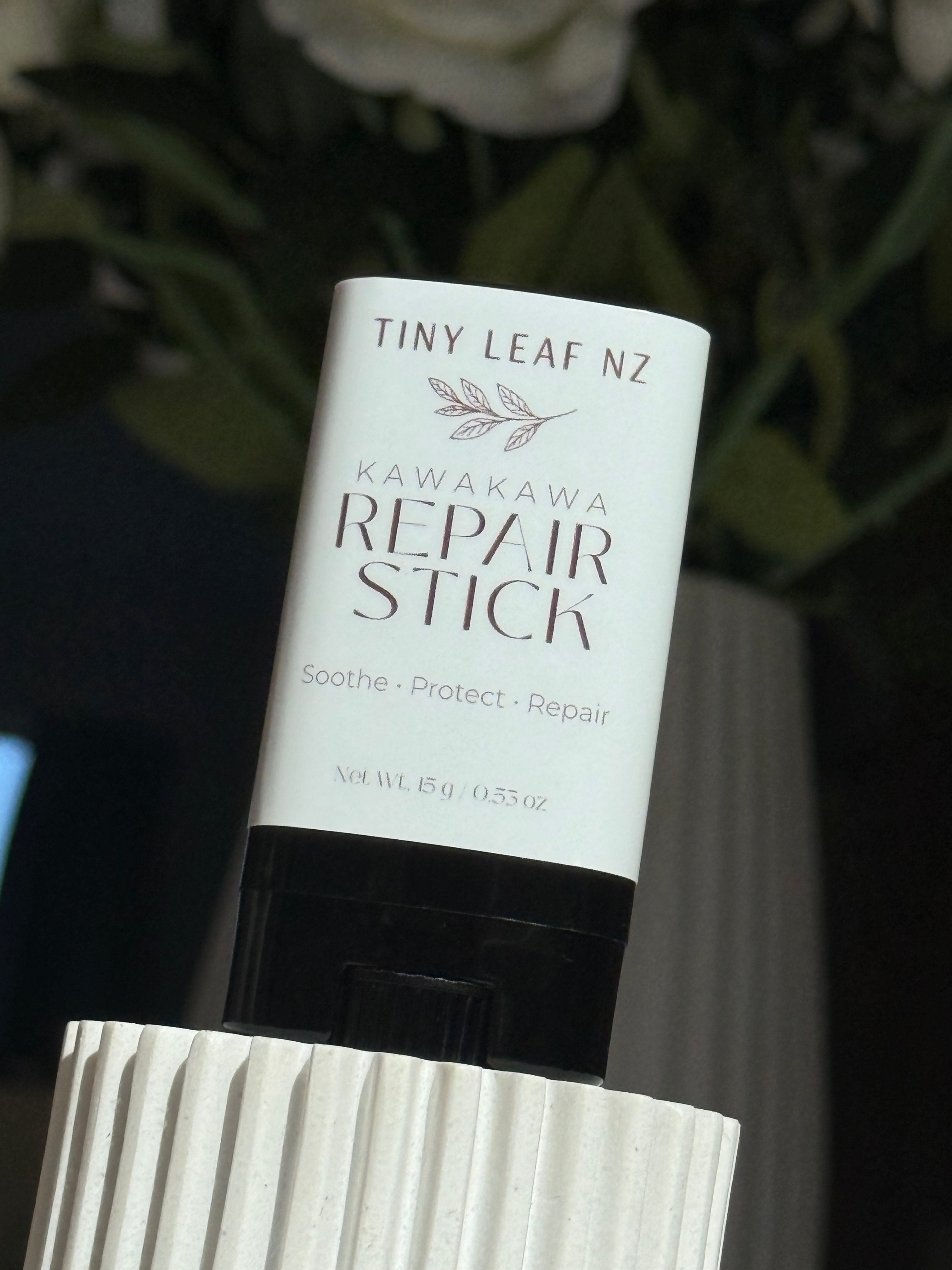 A white tube of Tiny Leaf NZ Kawakawa Repair Stick with a black cap, displayed on a white ribbed pedestal against a dark, blurred background with green leaves.