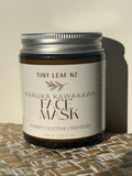 A jar of Tiny Leaf NZ Manuka Kawakawa Face Mask sits on a woven straw mat against a plain beige wall, casting a strong shadow to the right.