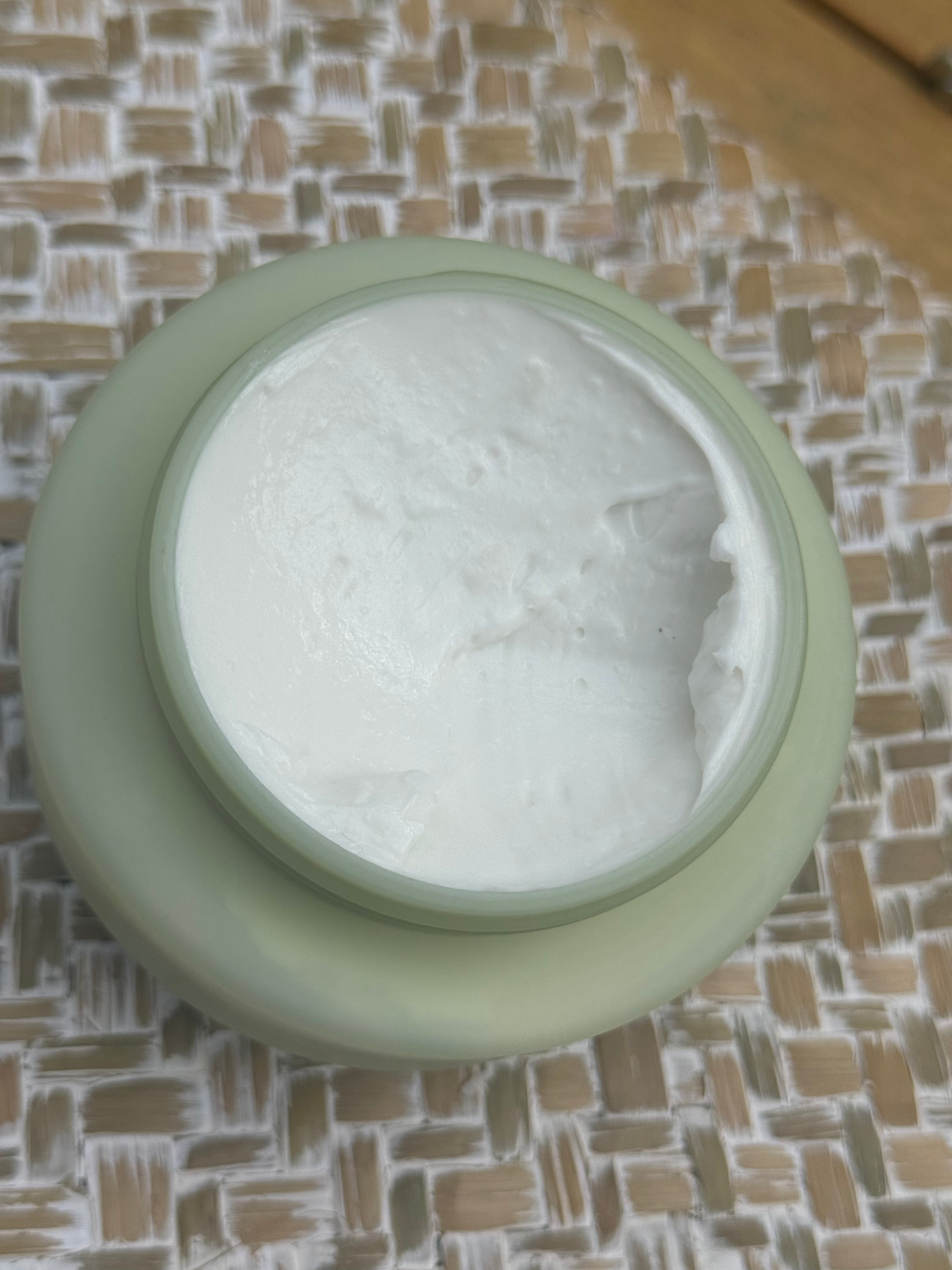 An open round cosmetic jar filled with white cream, placed on a woven beige and white textured mat.