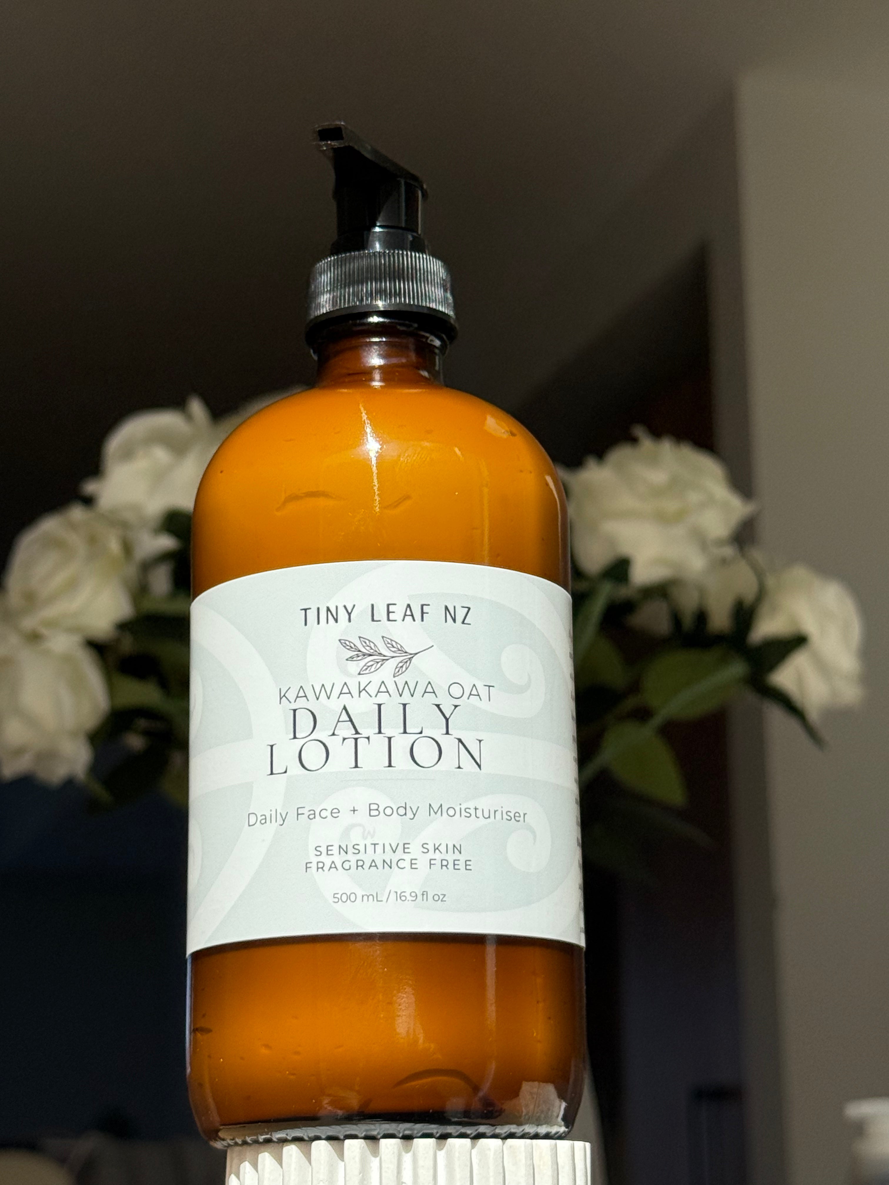 A bottle of Tiny Leaf NZ Kawakawa Oatmeal Daily Lotion with an amber-colored bottle, white label, and black pump dispenser, set against a blurred background with white roses.