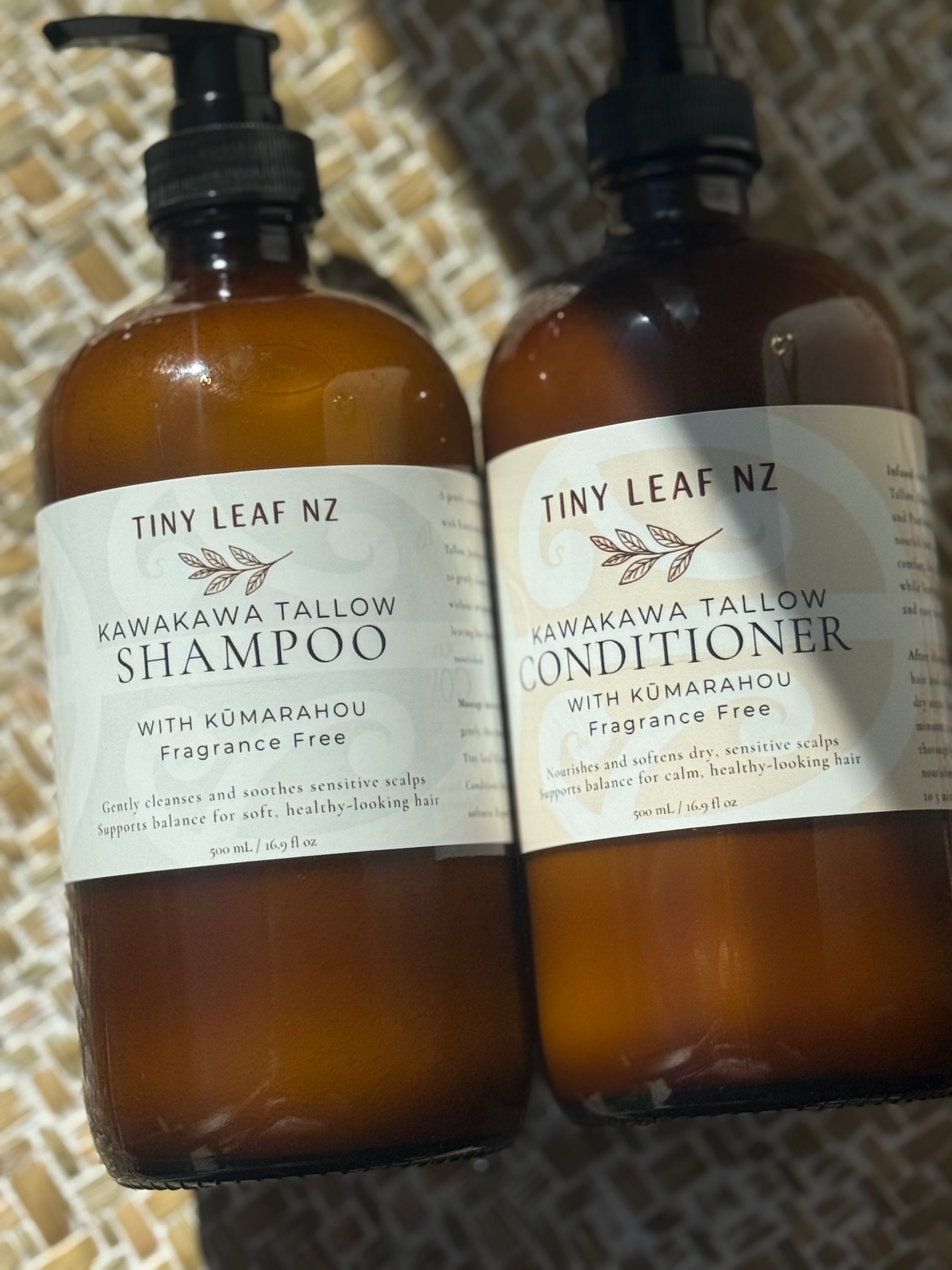 Two amber glass bottles of Tiny Leaf Nz hair care products, labeled Kawakawa Tallow Shampoo and Kawakawa Tallow Conditioner, with black pump and cap tops, set against a woven tan background.