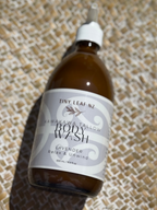 A bottle of Tiny Leaf Nz Lavender Body Wash resting on a woven straw mat. The bottle is amber brown with a white dropper cap and a white label featuring black and gray text.